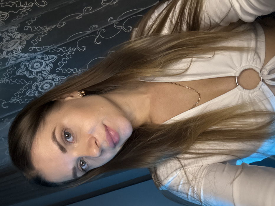 A woman with long blonde hair, wearing a white top with circular accents, poses for a photo. She has fair skin, light eyes, and is wearing a small earring. A gold chain necklace is visible. The background appears to be a patterned fabric, likely a bedspread. The image is lit in a way that creates highlights on her face and hair, while the background remains somewhat darker.