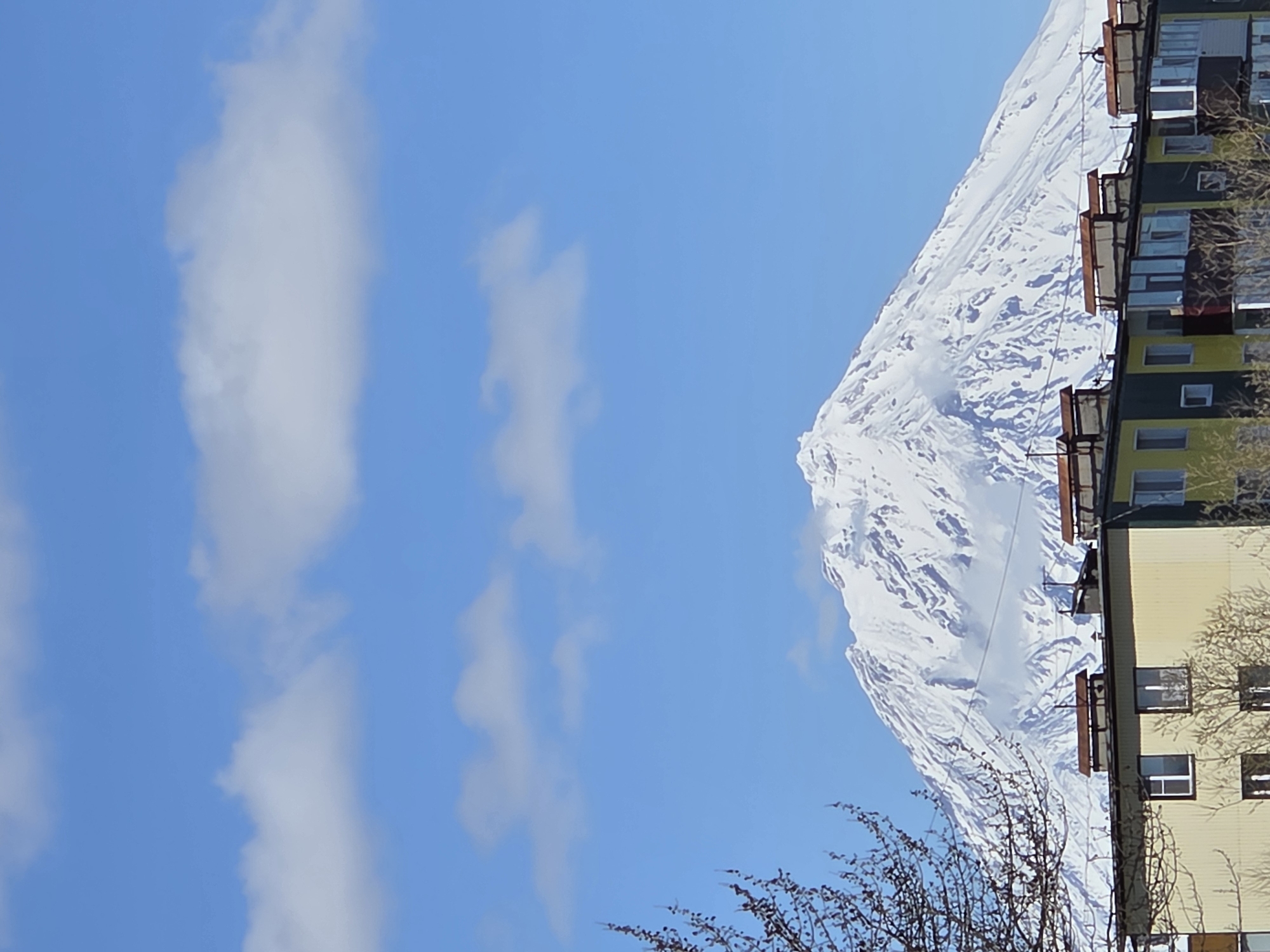 The image shows a building in the city of Unknown, Unknown, with a snow-covered mountain in the background. The building is multi-story, with multiple windows and balconies. The mountain is large and dominates the background. There are also some trees in the foreground. The sky is blue with some clouds.
