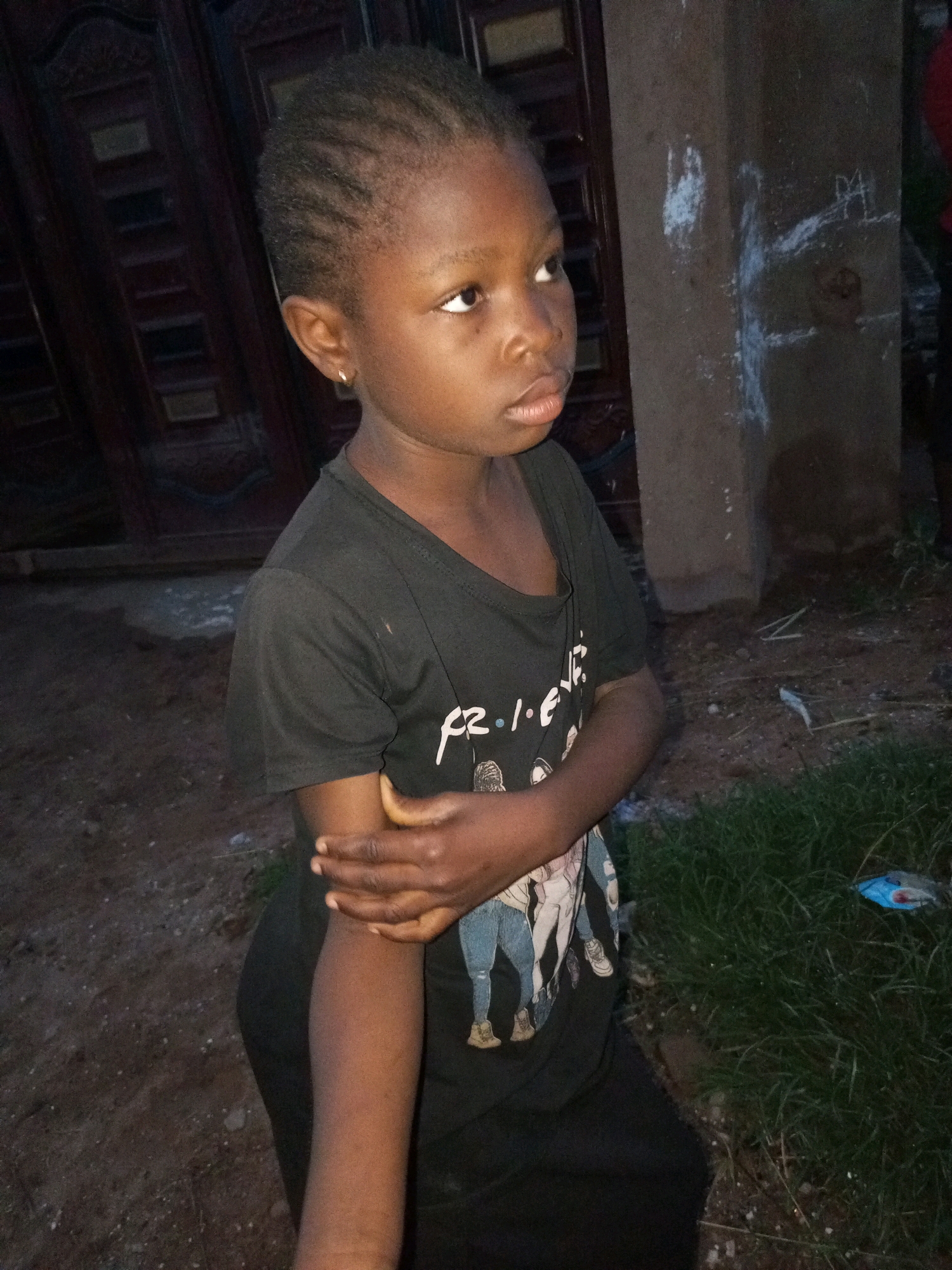 A young Black girl, with braided hair and wearing a black t-shirt with a cartoon graphic, stands in an outdoor area. She is looking to her right, with her left arm bent across her stomach. The background features a dark wooden door on the left and a concrete pillar with white markings on the right. The ground is dirt with some patches of grass and scattered debris. The image was taken in Tempure, Nigeria.