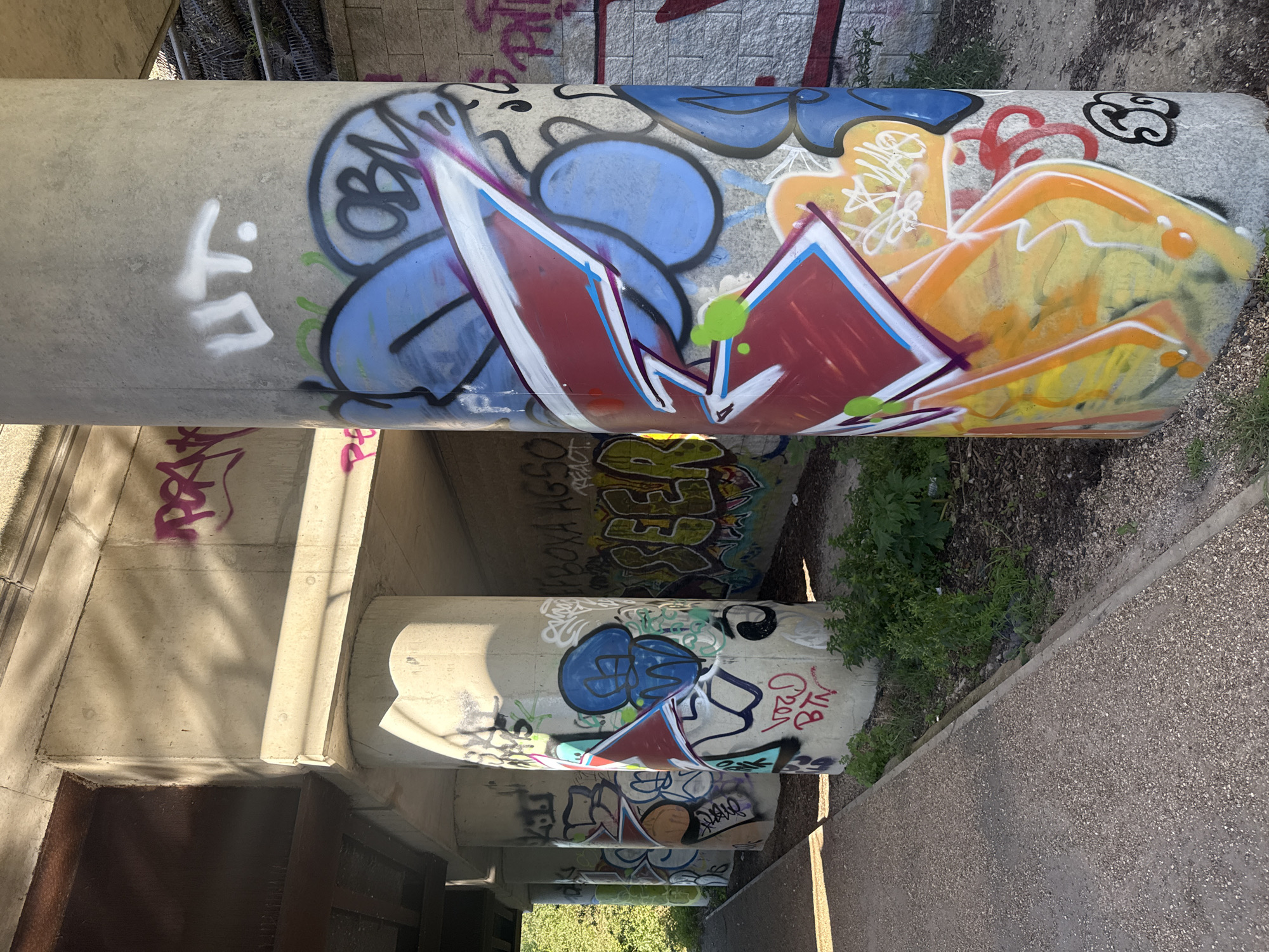 The media file captures the underside of a concrete bridge or overpass structure, likely in an urban setting in London, United Kingdom. The image is oriented sideways, presenting multiple concrete support pillars that span horizontally across the frame, all extensively adorned with colorful graffiti.
The most prominent feature is the large, light grey concrete pillar in the upper part of the image. On its left, a distinct white tag spells "LIT." Moving right, a blue bubble-style graffiti piece, possibly reading "OBM," is outlined in black. This is followed by a striking geometric design in red, white, and blue, highlighted with purple and green details. Further along the pillar, abstract yellow and orange graffiti with white outlines and black swirls adds to the vibrant display.
Beneath the main pillar, other concrete supports are visible, partially veiled by shadows. These too are covered in various tags and pieces. Recognizable text includes red letters spelling "NYA" and "BTN," alongside a clear green tag that appears to read "GREED." The overall impression is one of dynamic, unauthorized artistic expression transforming the dull concrete environment.
To the right of the pillars, the ground is visible, composed of gravel, dirt, and scattered green weeds and plants. Strong sunlight creates sharp shadows across the scene, indicating a clear and sunny day, possibly morning or afternoon. The concrete surfaces exhibit textures and some accumulated grime, typical of an overlooked urban space, which serves as a canvas for the street art.