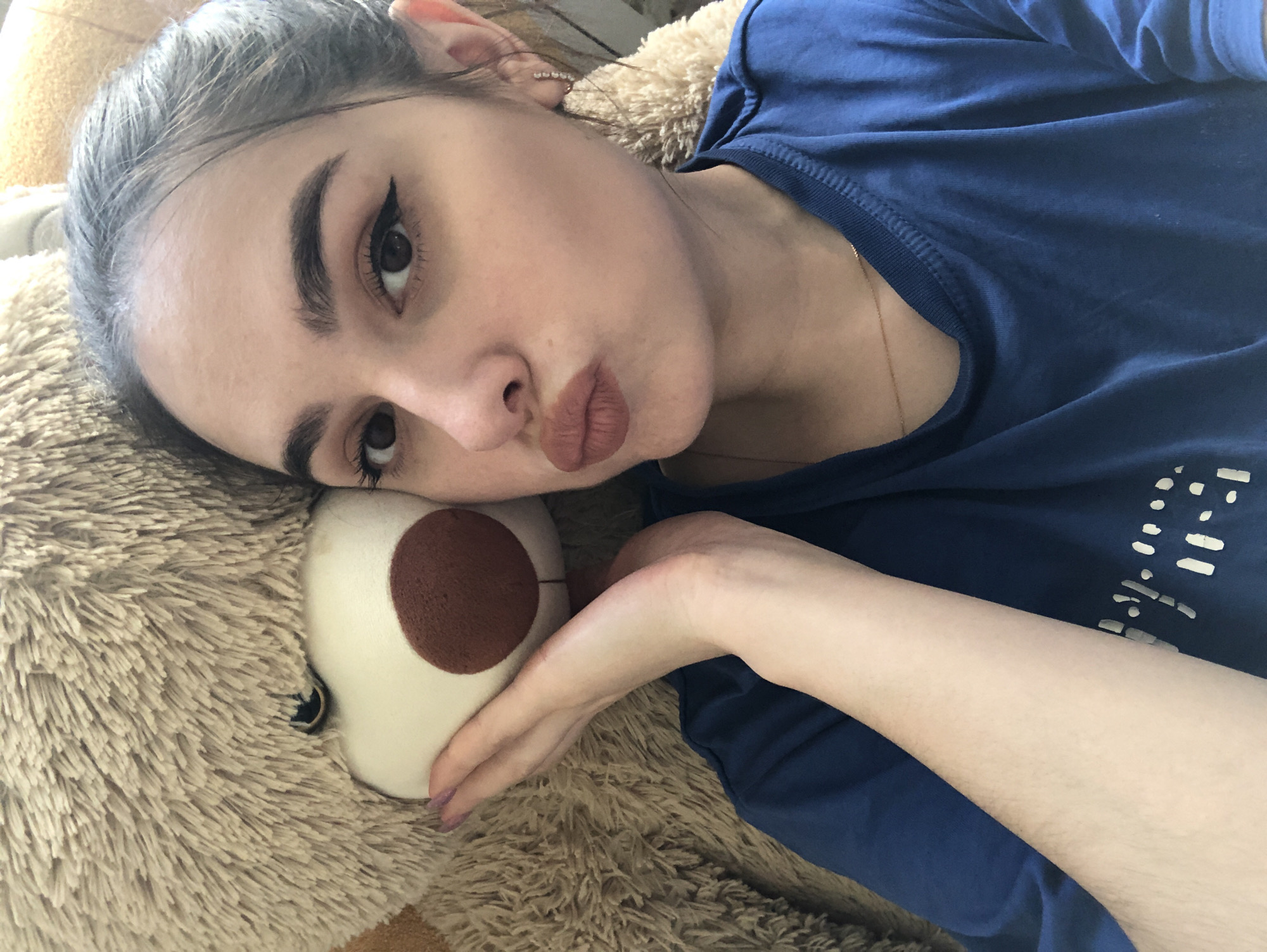 A woman is lying next to a teddy bear. The woman has her hair pulled back, and she is wearing eyeliner and lipstick. She is looking at the camera with her lips pursed. She is wearing a blue shirt. The teddy bear is large and furry, with a brown nose. The background is out of focus. There is no information in the image to indicate a location in Noviy, Russia.