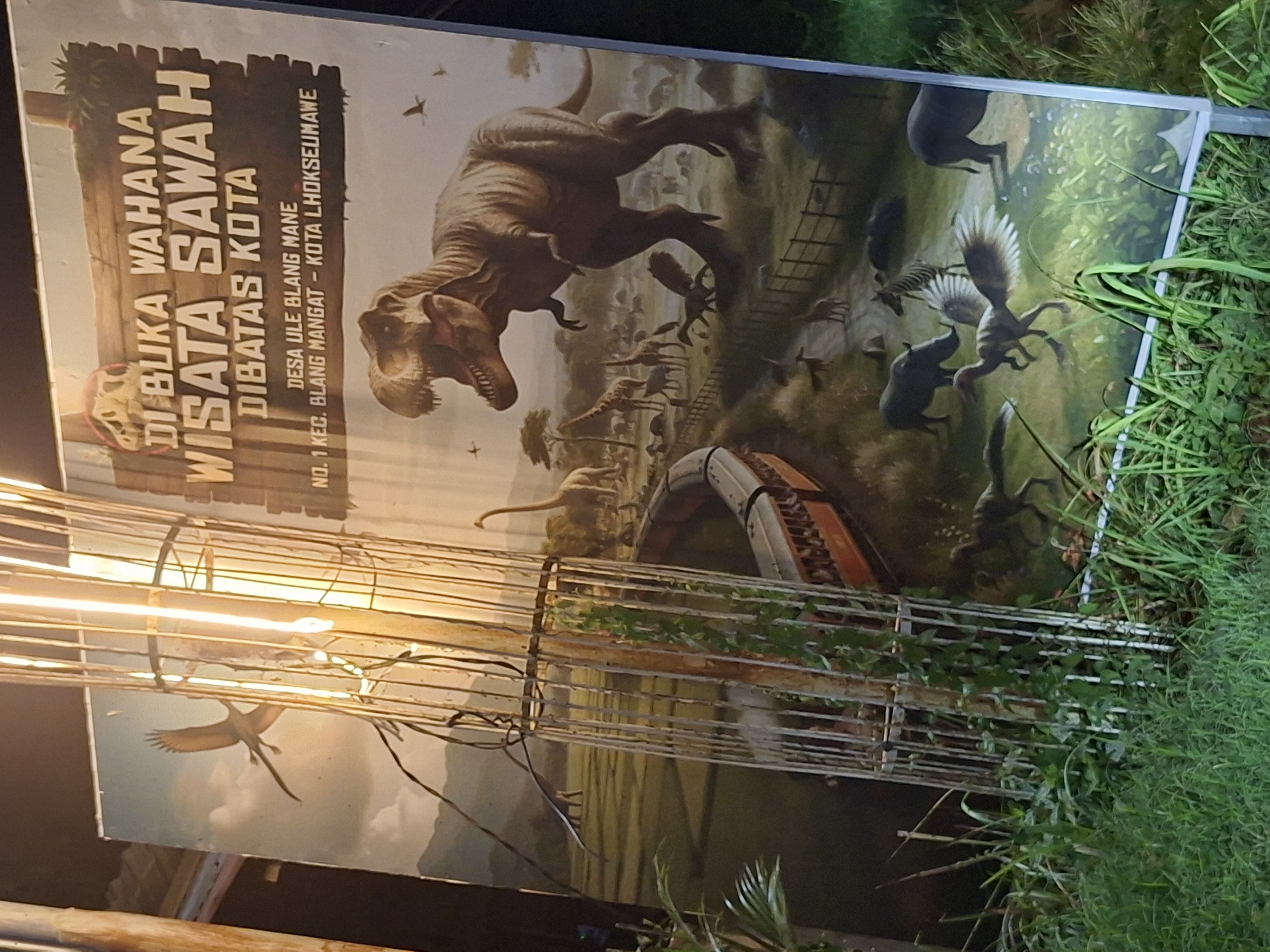The image shows a sign in Indonesian that reads "DI BUKA WAHANA WISATA SAWAH DIBATAS KOTA, DESA ULE BLANG MANE, NO. 1 KEC. BLANG MANGAT - KOTA LHOKSEUMAWE." This translates to "Opening Tourist Rice Field Attraction, Bordered by the City, Ule Blang Mane Village, No. 1 Blang Mangat District - Lhokseumawe City." The sign features illustrations of dinosaurs, giving the impression of a dinosaur-themed attraction near a rice field within or bordering Lhokseumawe, Indonesia. The sign is placed near some foliage, including what looks like grass. A decorative metal cage-like structure partially obscures the lower portion of the sign.