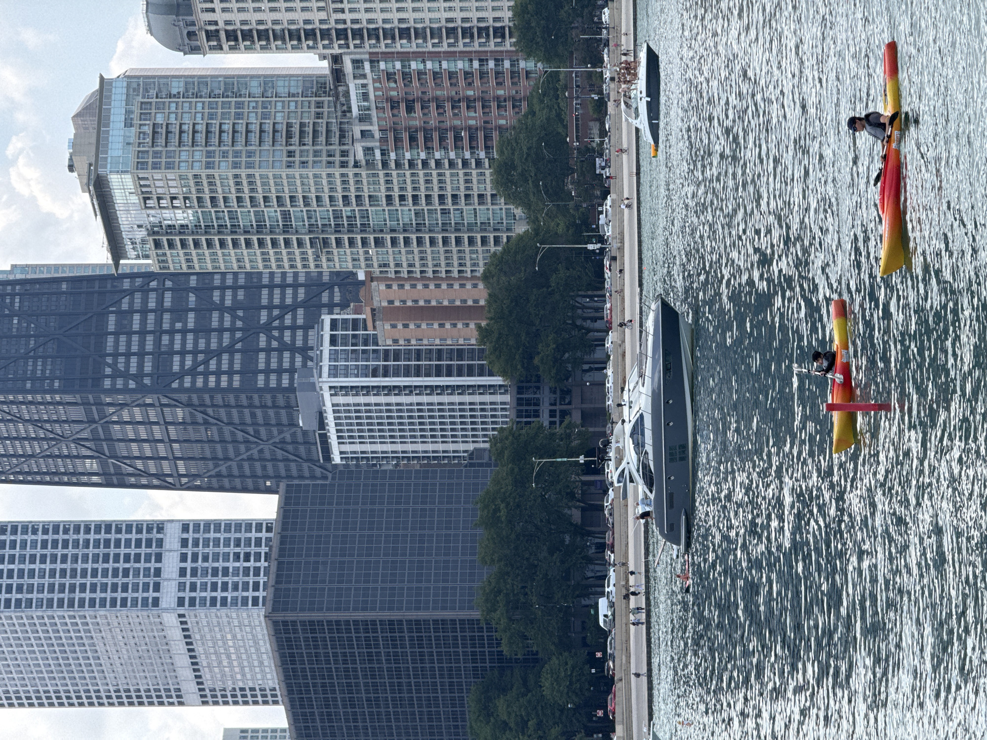 The image depicts a scene along the Chicago waterfront, featuring a backdrop of the city's iconic skyline. Dominating the background are several tall buildings, including a distinctive black skyscraper with a lattice-like exterior, and a lighter colored, grid-patterned skyscraper. In the mid-ground, trees line a walkway, with a street visible behind them populated by cars and pedestrians.
In the foreground, the rippling surface of Lake Michigan occupies the majority of the frame. Two individuals are visible in kayaks. The kayak in the foreground is yellow and red, with one person wearing a dark cap and t-shirt, paddling with a black oar. Behind this, another yellow kayak is visible with one person paddling. A red marker buoy floats in the water between the two kayaks.
Further out on the water, two motorboats are present. The larger of the two, a sleek, dark-colored yacht, is positioned on the left side of the frame. On the right, a white motorboat is visible with several individuals standing on its deck. The overall scene suggests a warm, possibly summer day, with recreational activities taking place on the water against the urban landscape.