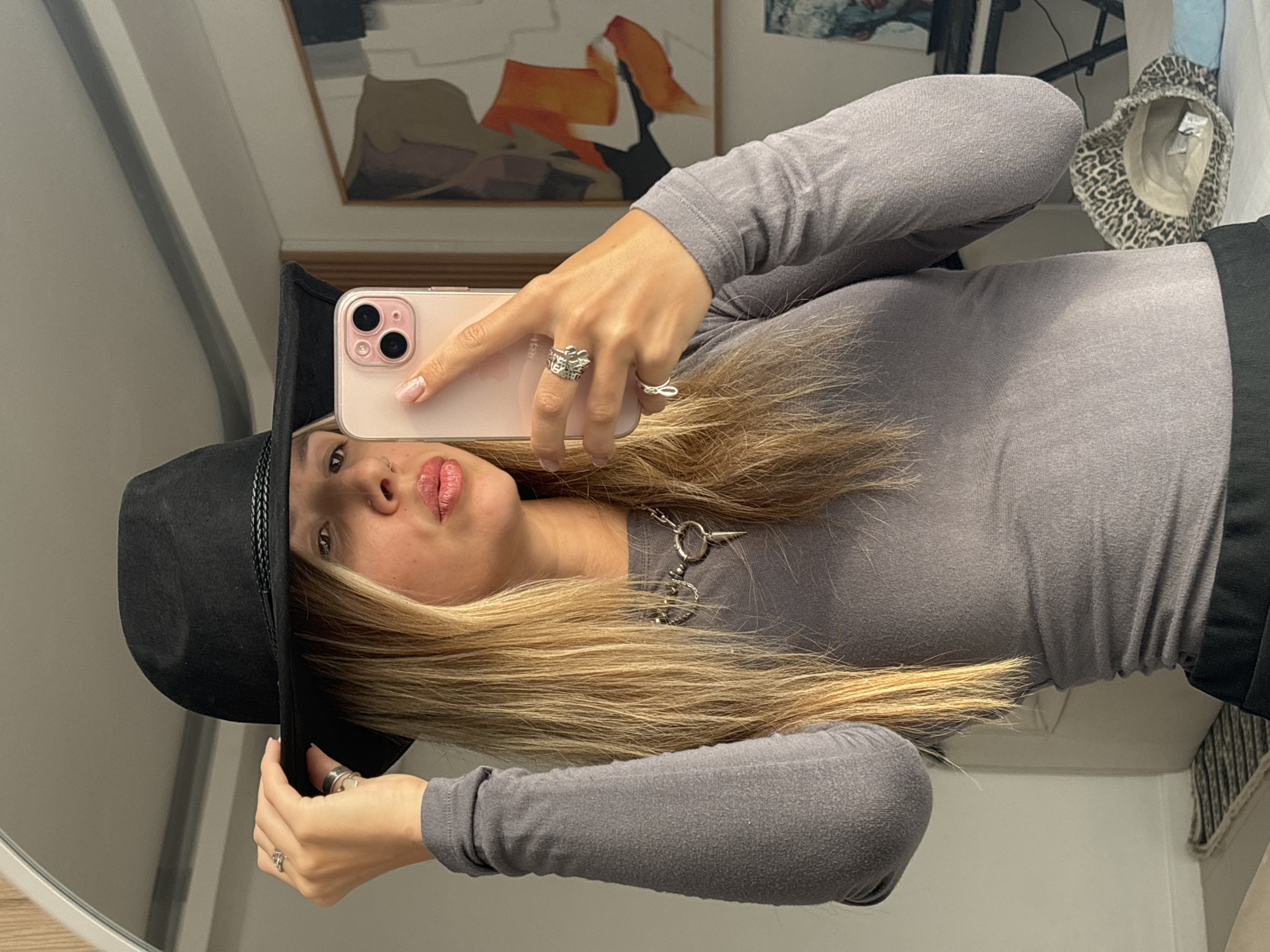 A young woman is taking a mirror selfie in a bedroom. She is wearing a grey long-sleeved shirt, a black hat, and a silver necklace. She is also wearing rings on her fingers and has blonde hair. In the background, there is a painting on the wall and a keyboard. The room appears to be well-lit, and the overall atmosphere is casual.