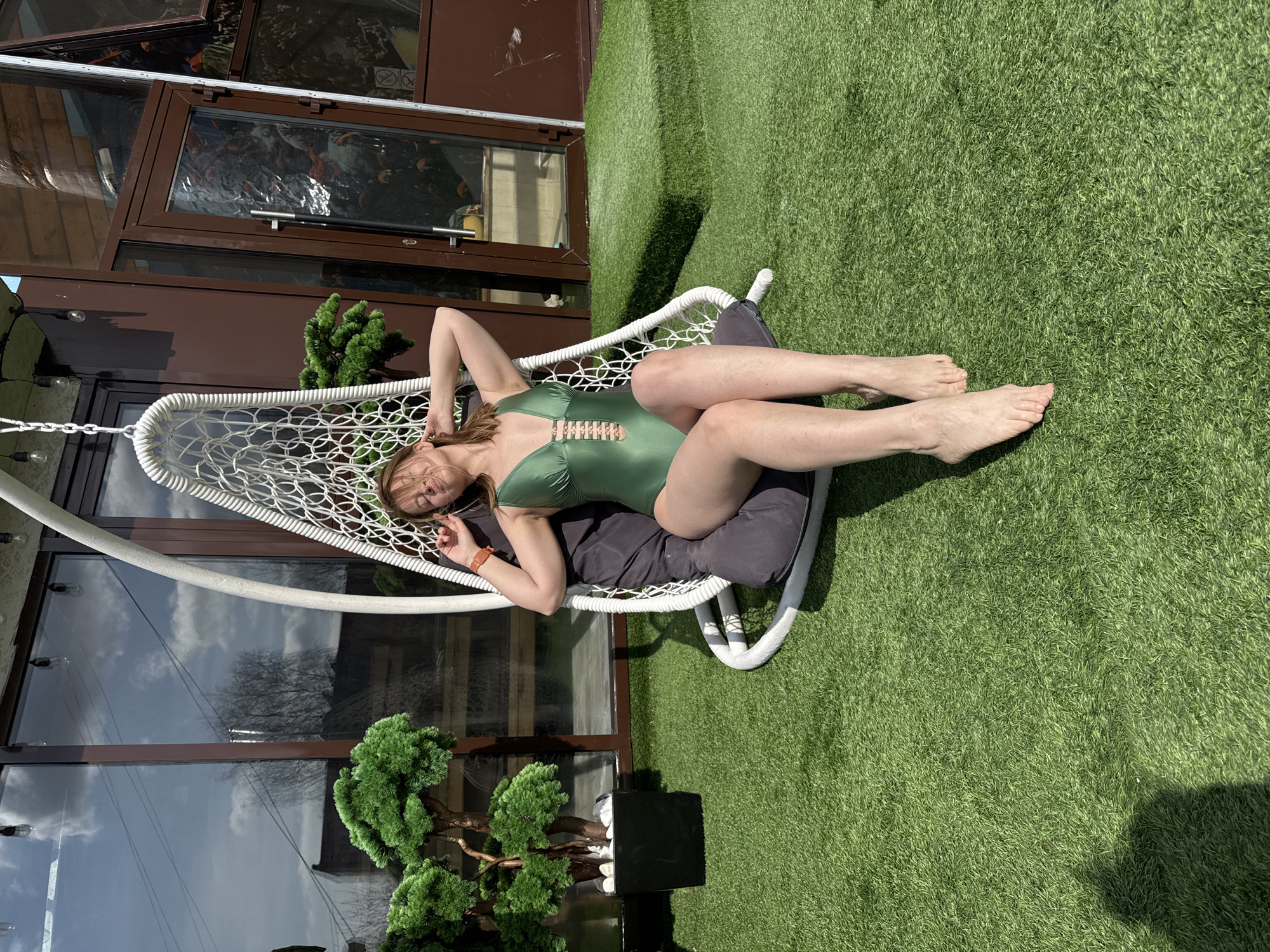 A woman in a green swimsuit reclines in a white hammock chair on a green lawn. The background includes a building with brown window frames, reflecting trees and sky. A decorative bonsai tree stands near the building. The location is likely a private yard or patio in Votkinsk, Russia, indicated by the context provided.