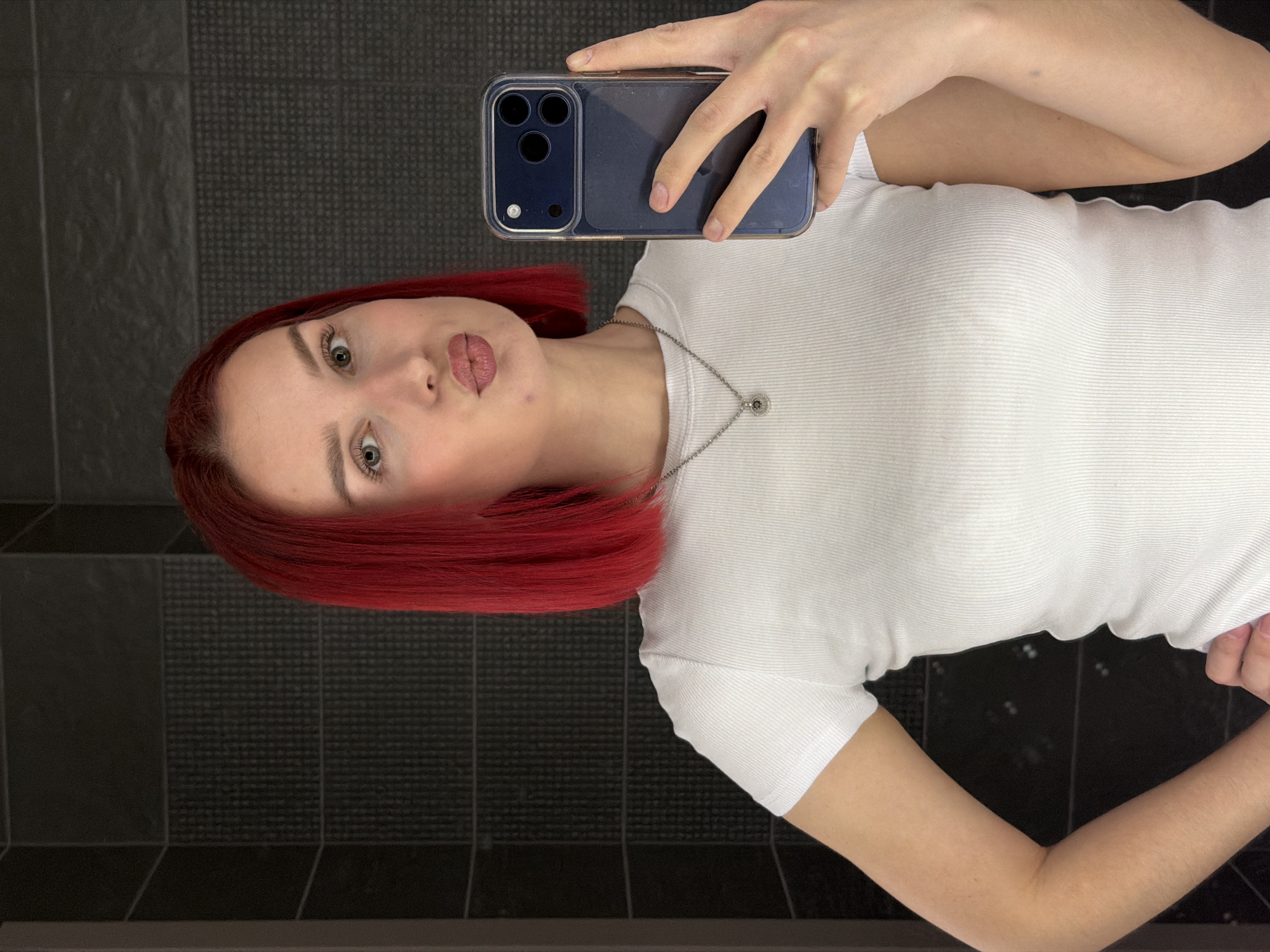 A young woman with short, bright red hair is taking a selfie in what appears to be a bathroom. She is wearing a white ribbed t-shirt and a delicate silver necklace. She is pouting slightly at the camera. The phone she is using to take the picture is blue with a clear case. The background is a dark gray tiled wall. The image was taken indoors. There's no indication of location in the city of Chaikovsky, Russia.
