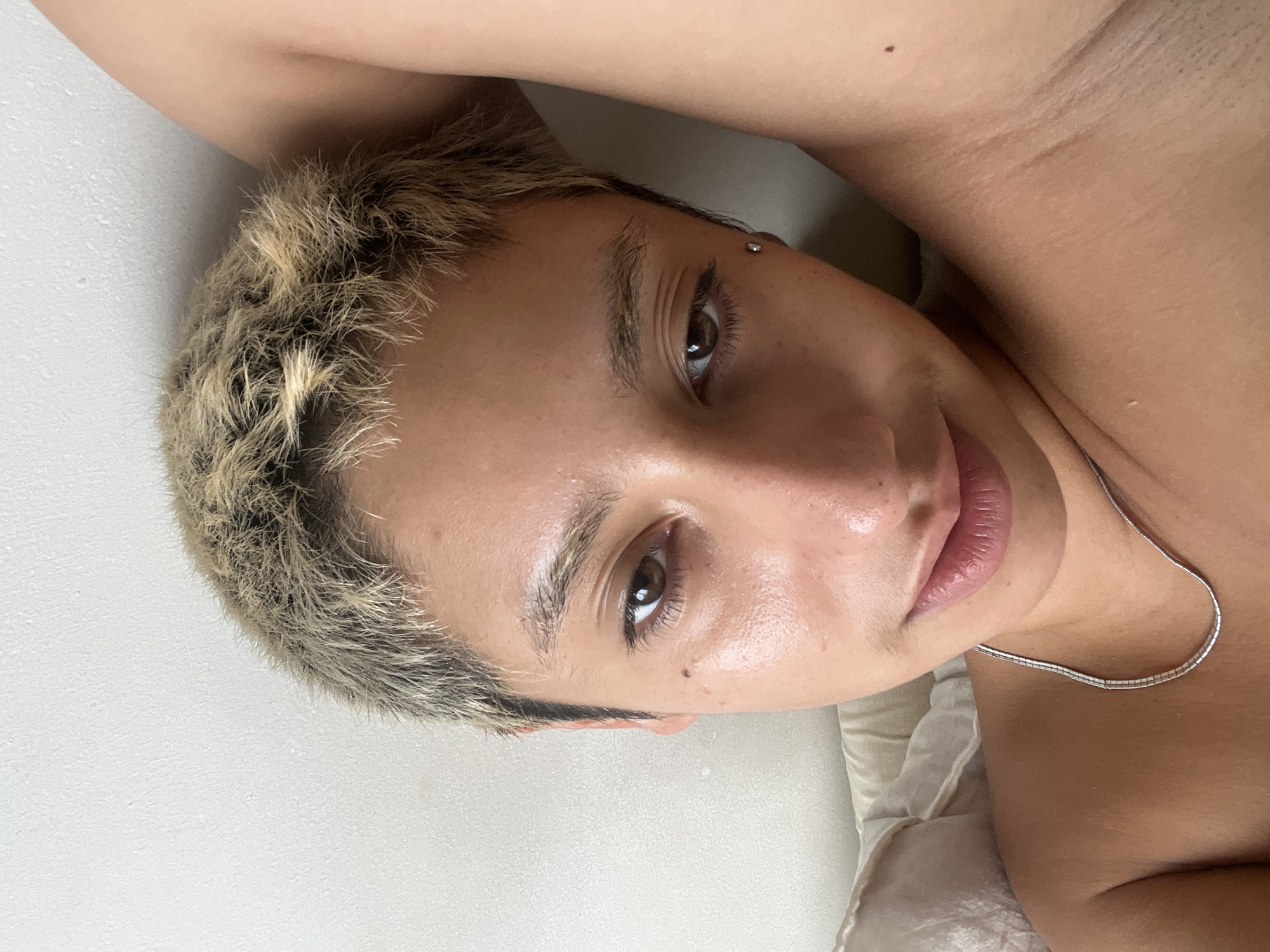 A close-up, slightly angled portrait of a person with light skin, light brown eyes, and short, textured hair dyed in a blonde and brown color combination. The person is reclining on what appears to be a light-colored surface, possibly bedding. A thin, silver necklace is visible on their neck. There is a piercing visible near the upper brow line. No other individuals are visible in the frame, and the setting is indoors. There is no indication of the location being Buenos Aires, Argentina, within the image.