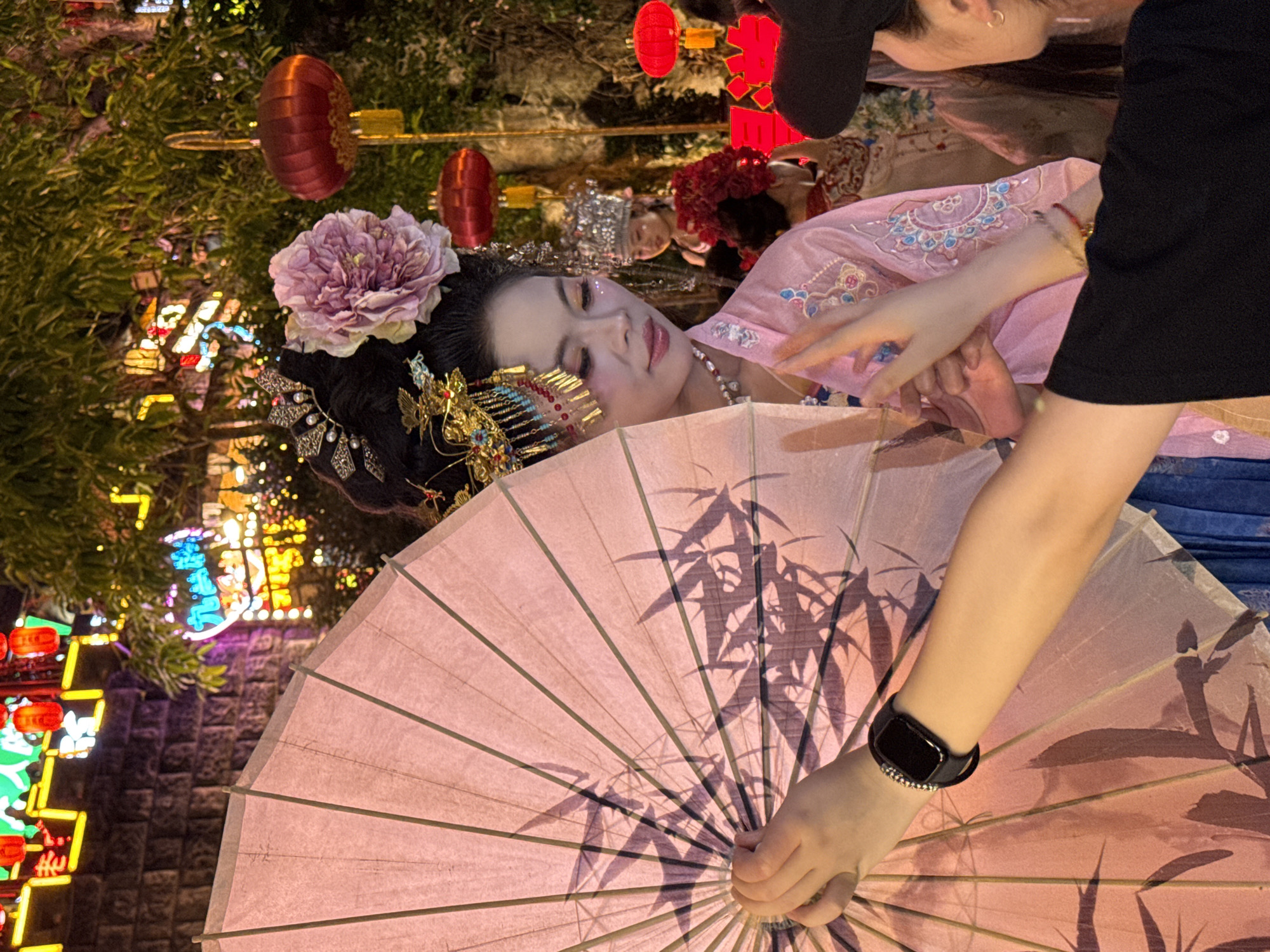 In Yuzhong District, China, a woman in traditional Chinese attire, complete with an ornate headpiece and a large pink parasol, is the central subject. She is adorned with elaborate makeup featuring glitter on her eyelids and cheeks. The parasol, decorated with a bamboo leaf motif, is held to her right, partially obscuring her face. Her left hand is visible, holding the parasol, and a smart watch is worn on her left wrist. She is wearing a pink, embroidered robe.
To her left, another person, dressed in a black t-shirt and a black baseball cap, is reaching out towards the woman with the parasol. This individual's right arm is extended, and their fingers are positioned near the woman's hand.
In the background, a festive scene unfolds. Red lanterns are hung, and neon lights, some in Chinese characters, illuminate the area. Trees with glowing lights are also visible, suggesting a night-time celebration or event. Other individuals are present in the background, also appearing to be dressed in traditional attire. The overall environment suggests a lively, possibly cultural, gathering.