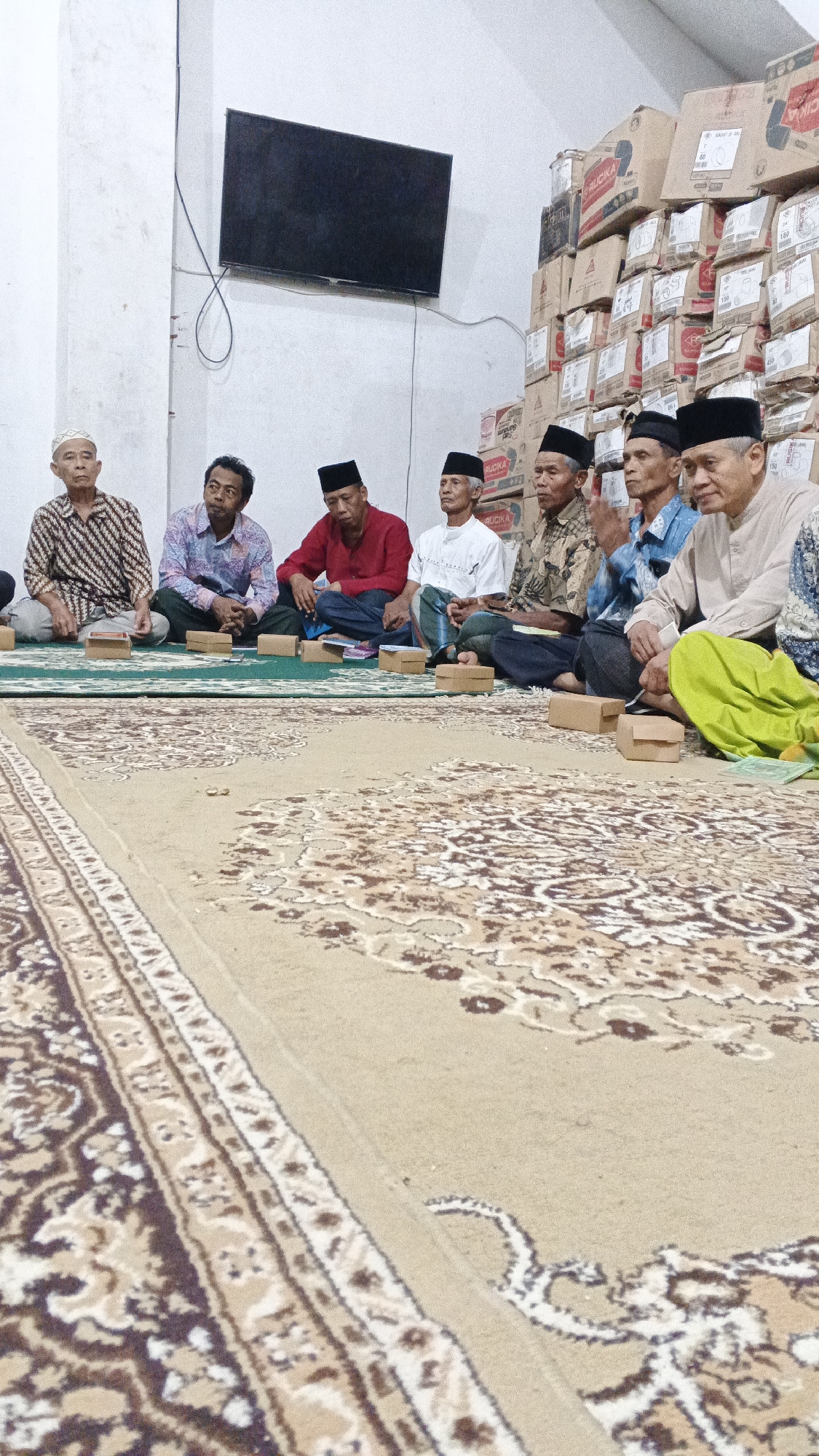 A group of nine elderly Indonesian men are seated cross-legged on a patterned rug inside a room. The room has white walls and a flat-screen television mounted on the left wall. Behind the men, stacked cardboard boxes are visible, some bearing the brand name "RUCKA". The men are dressed in traditional Islamic attire, including peci caps and long-sleeved shirts or robes. Some of the men are looking towards the camera, while others are looking down or at each other. Small, brown cardboard boxes are placed in front of some of the men. The scene appears to be a gathering or meeting in a modest indoor setting, possibly a community hall or home, located in Ambarketawang, Indonesia.
