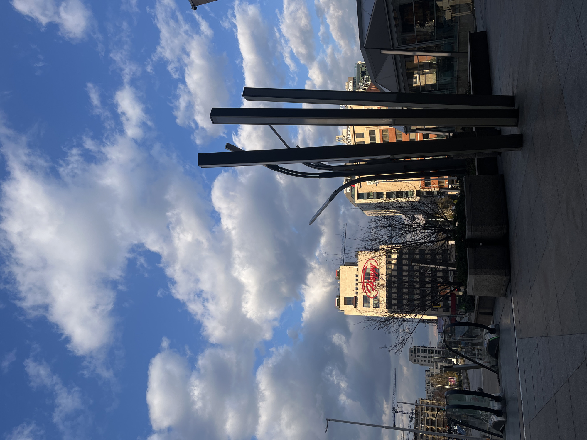 The image shows a street scene in Sofia, Bulgaria. The viewpoint is from a pedestrian area, with a view of a building bearing the "Ferrero" logo. Modern, dark-colored light poles/art installations are visible in the foreground. The sky is partly cloudy, with a mix of blue sky and white clouds. Other buildings and construction cranes are visible in the background. The photo is taken from a slightly low angle, giving prominence to the sky and the building with the logo.