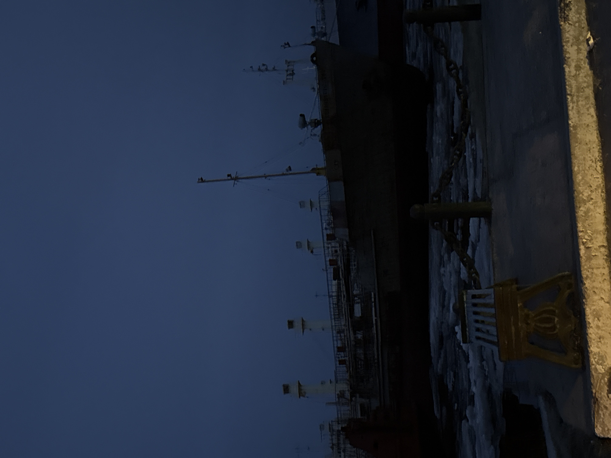 A long ship is docked, viewed from ground level at twilight. The ship is dark, with a complex superstructure and a tall mast. A concrete barrier is on the right, with a decorative gold mooring cleat. The sky is a uniform dark blue. Based on the presence of ships and docking facilities, the location might be in or near the city of Zhatay, Russia, given its location on the Lena River which hosts shipping activity. No people are visible.