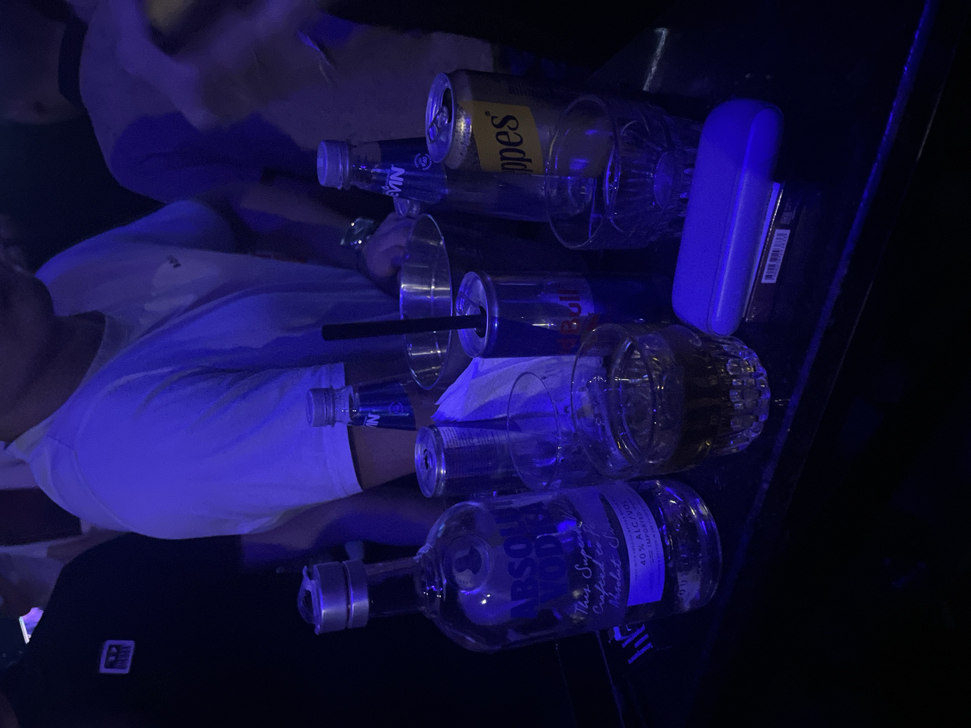 The image shows a bar scene, likely in Plovdiv, Bulgaria, with several people and drinks on a dark surface. Visible items include a bottle of Absolut Vodka, several drinking glasses (some empty, some filled with liquid), a can of Schweppes, a blue bottle of tonic, a black drinking straw, a blue eyeglasses case, and a small black item with a white square. People are partially visible, with one person wearing a white shirt. The scene is lit with blue lighting.