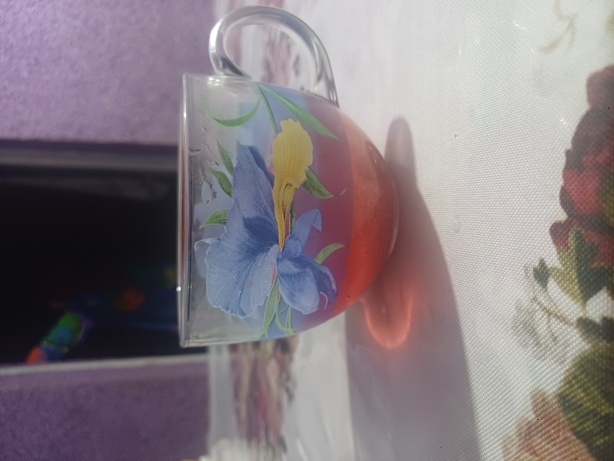 A clear glass mug decorated with a blue and yellow flower design sits on a white tablecloth with a floral pattern. The mug contains a red liquid. The background is out of focus and shows a purple wall and a dark vertical object with colorful reflections. The scene is lit by a light source from the left, casting a shadow of the mug on the tablecloth.