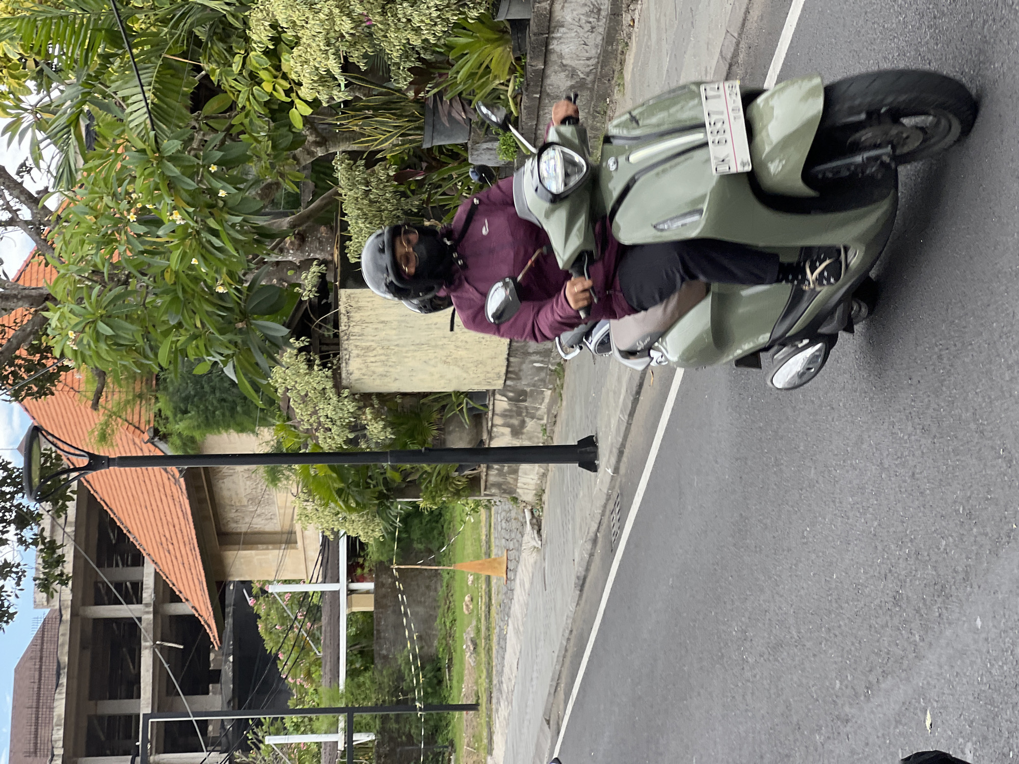 In Denpasar, Indonesia, a person wearing a maroon jacket, black helmet, and face mask rides a light green scooter on an asphalt road. The scooter has a license plate that reads "DK 6657 TZ". In the background, there is a building under construction with exposed concrete beams and an orange tiled roof. Lush green trees and plants line the sidewalk, and a black lamppost stands near the road. The sky is overcast.
