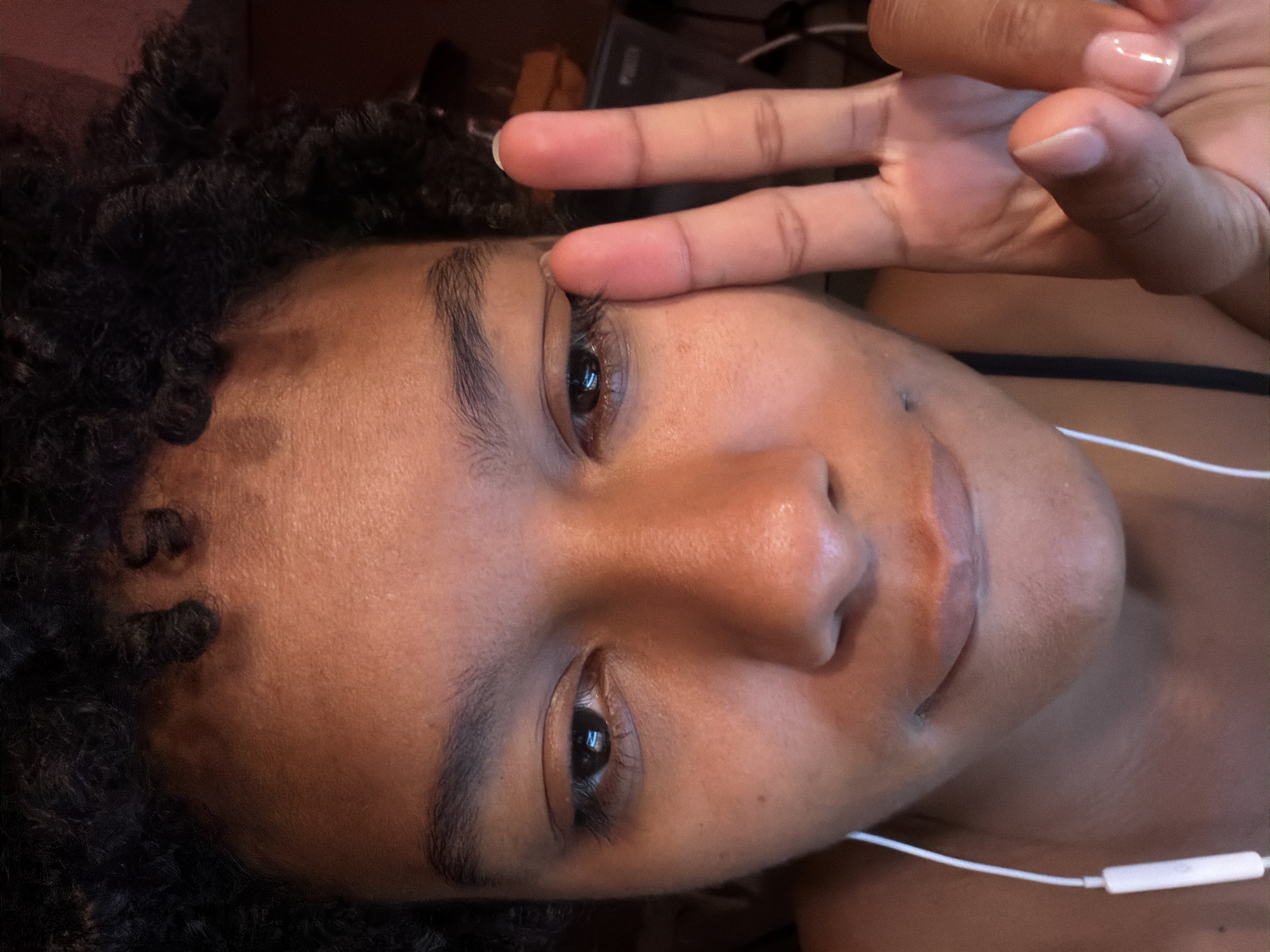 A person with curly black hair and brown skin is angled with their head near the top left of the frame. They are smiling with a hand raised in a peace sign, one finger resting on their eyelid. A white wired headphone is positioned to the right, trailing down the neck. The background is dark and indistinct, with a glimpse of a light-colored object near the top center. The photo was not taken in Champs-sur-Marne, France.