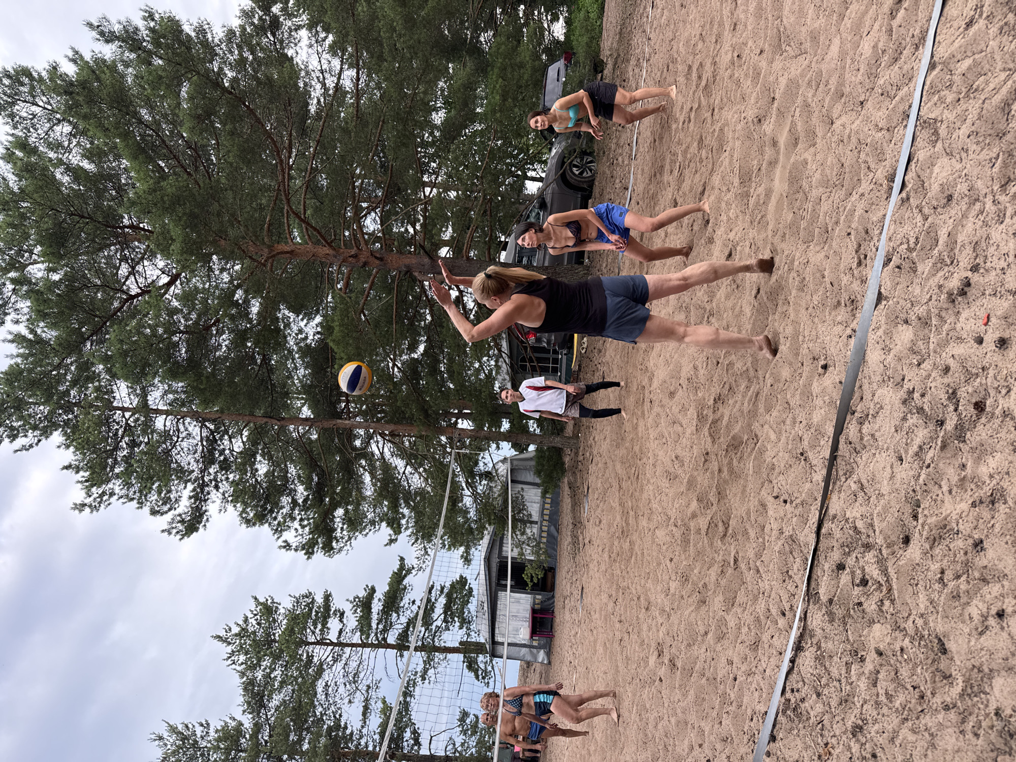 A group of six adults are playing beach volleyball on a sand court. Two people are on one side of the net, and four are on the other. A woman in a black tank top and blue shorts is in the process of serving the ball, which is in the air above her. Another woman in a blue bikini top and black shorts is positioned to receive the serve. The background shows pine trees, a small building, and two cars. The sky is overcast. The image appears to have been taken in Zelenaya Roshcha, Russia.