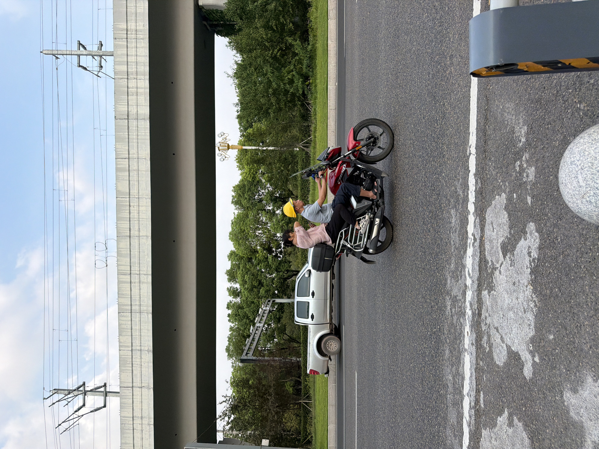 A daytime, eye-level, medium shot captures a motorcycle with two occupants traveling on an asphalt road in Jiangjin, China, beneath a concrete overpass with overhead electrical lines. The motorcycle, a red sport-style bike, is being driven by a man wearing a blue shirt and a yellow hard hat. Seated behind him is a woman in a pink and white striped shirt. A black storage box is attached to the rear of the motorcycle. To the left of the motorcycle, a silver pickup truck, also in motion, is visible. The background consists of lush green trees lining the road and a gray concrete overpass structure overhead. The road has white lane markings.