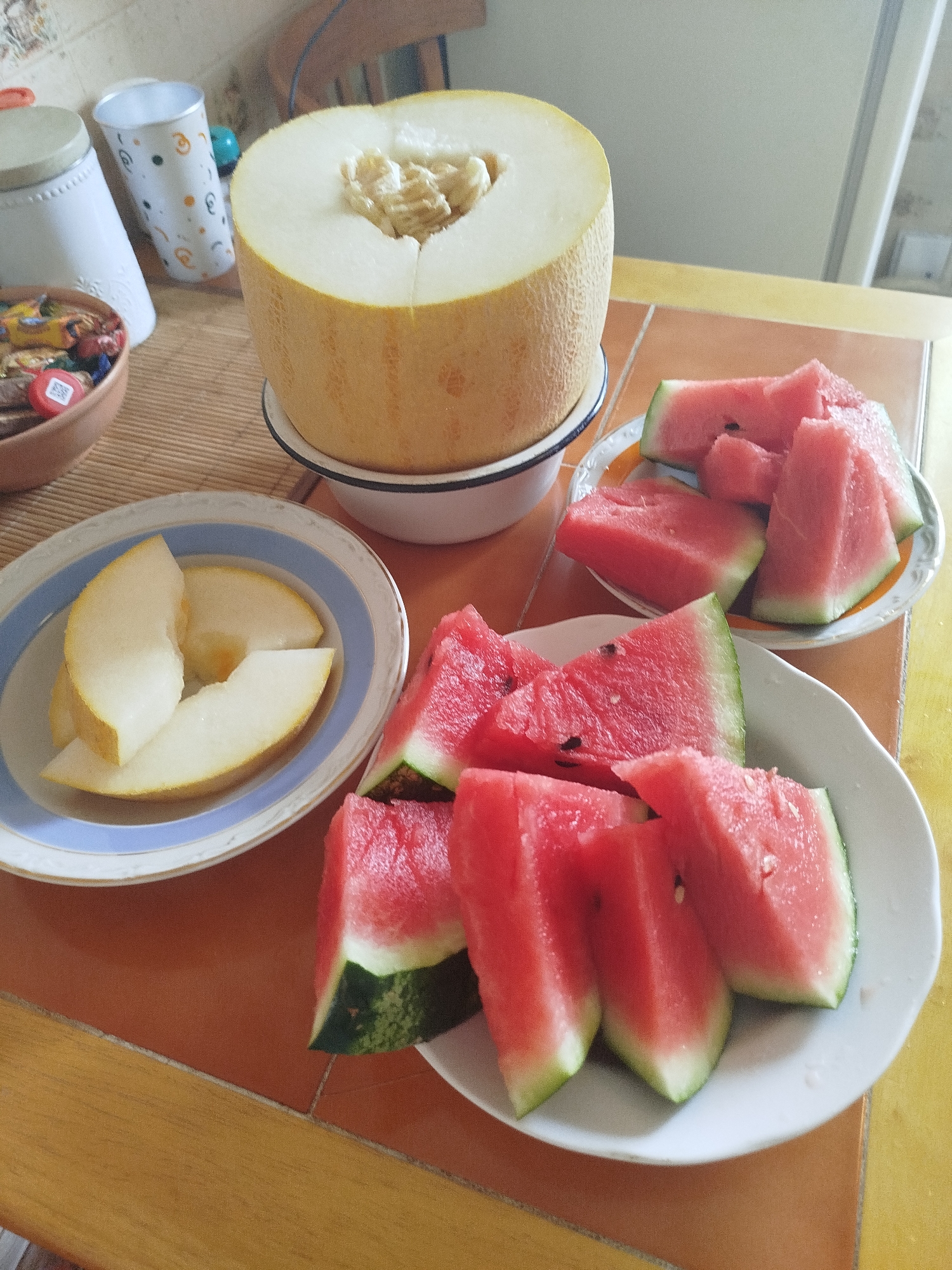 The image depicts a dining table with several fruits and other items. In the foreground, a white bowl contains several slices of watermelon. To the left of this bowl, a plate holds more watermelon slices. Behind these, a larger white bowl contains slices of melon. Further back, to the left, is a decorative white container and a patterned cup. To the right, a portion of a chair is visible. The table surface appears to be made of orange tile with a wooden edge. The scene is set indoors, likely in a kitchen or dining area. There are no people visible in the image. The lighting suggests it is daytime.