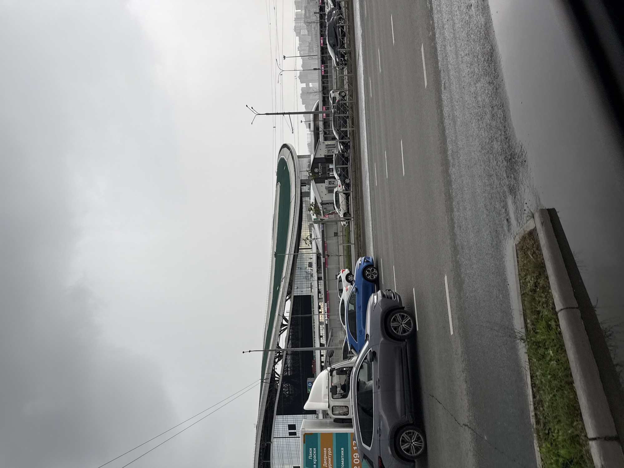 This image depicts an outdoor scene in Kazan, Russia, under overcast, possibly rainy, weather conditions. A multi-lane road runs along the right side of the frame, with its surface appearing wet and reflective. Several cars are parked or driving on the road, including a dark grey SUV, a white truck, a blue car, and a white car. The vehicles are positioned in a line parallel to the road.
To the left of the road, a large modern building with a curved green roof is visible. This structure appears to be a sports arena or entertainment complex, with the text "AK BARS ARENA" partially visible on its facade. The area in front of the building has a sidewalk and some grassy areas. Overhead power lines crisscross the sky. In the distance, a city skyline with numerous buildings can be seen under the grey sky.
The time of day is not definitively clear, but the dull lighting suggests it could be daytime under cloud cover, or possibly dusk. There are no visible people in the image. The overall mood is somewhat subdued, consistent with the cloudy weather. Visible text includes "AK BARS ARENA" and some Russian text on signage on the white truck, which appears to be advertising services related to paints and coatings ("Лаки и краски", "Дверная фурнитура", "Автоматика").