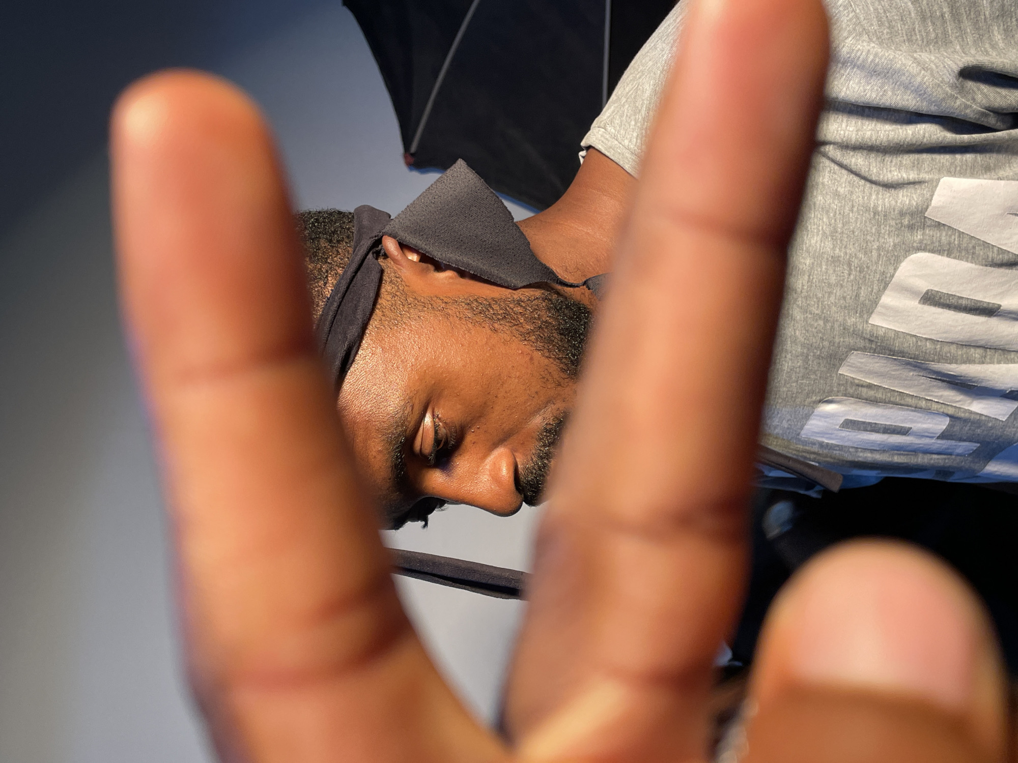 A close-up, slightly rotated image shows a dark-skinned person with a beard and a black head covering. The person's eyes are closed. Two out-of-focus fingers fill the foreground, obscuring parts of the subject. The person is wearing a grey t-shirt with white lettering. A black umbrella is visible in the background. The location is Barakin Akawo, Nigeria.