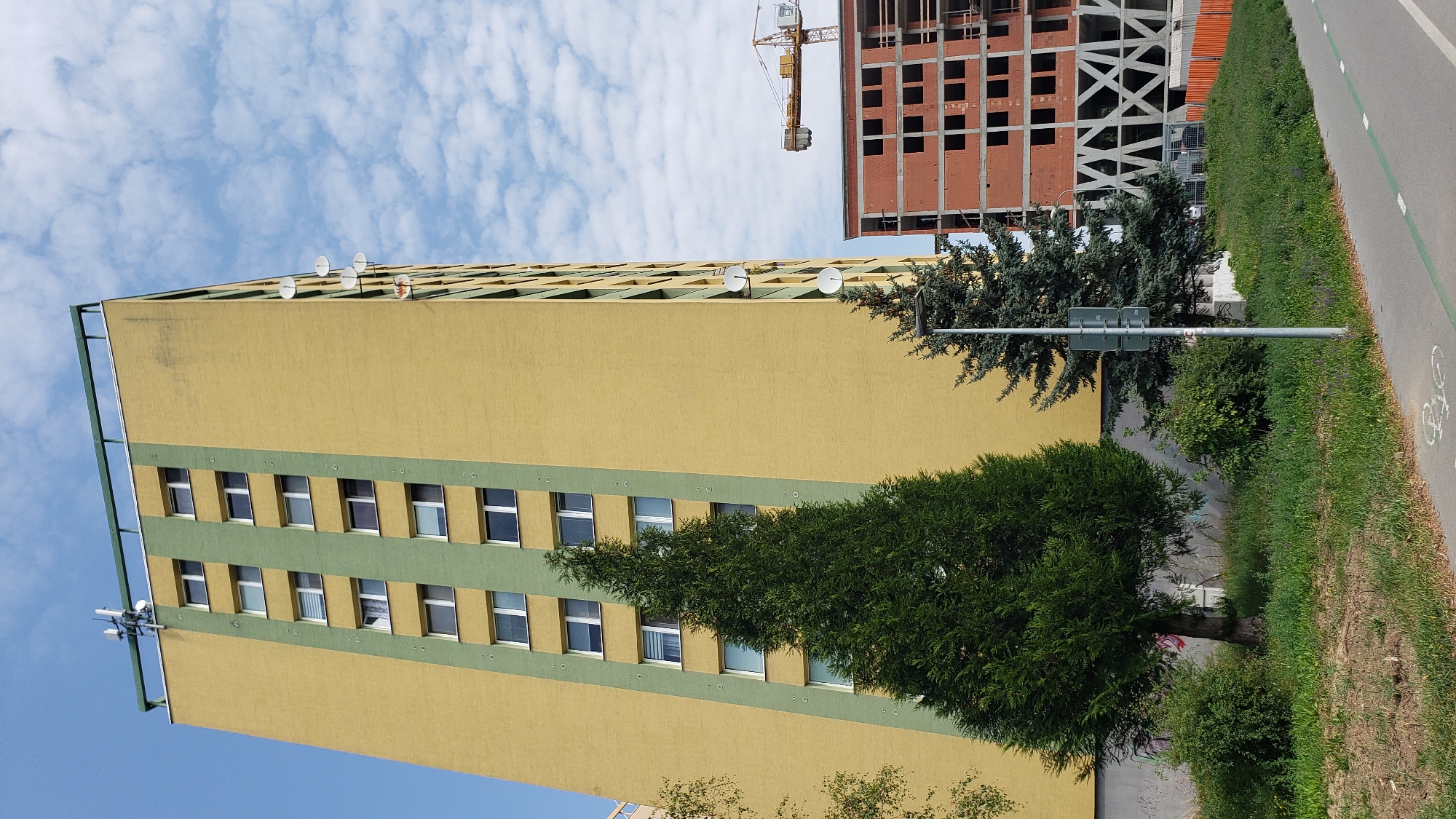 An exterior, low-angle shot captures a tall, yellow apartment building with green vertical accents, a large evergreen tree in the foreground, and a partially constructed brick building with a yellow crane in the background. The apartment building has multiple windows and several satellite dishes mounted on its side. The evergreen tree is dense and obscures part of the building. The unfinished building to the right is made of red brick and has exposed structural beams. The scene is set under a partly cloudy blue sky. This image was taken on Vihorlatská Street in Prešov, Slovakia. There are no people visible in the image.