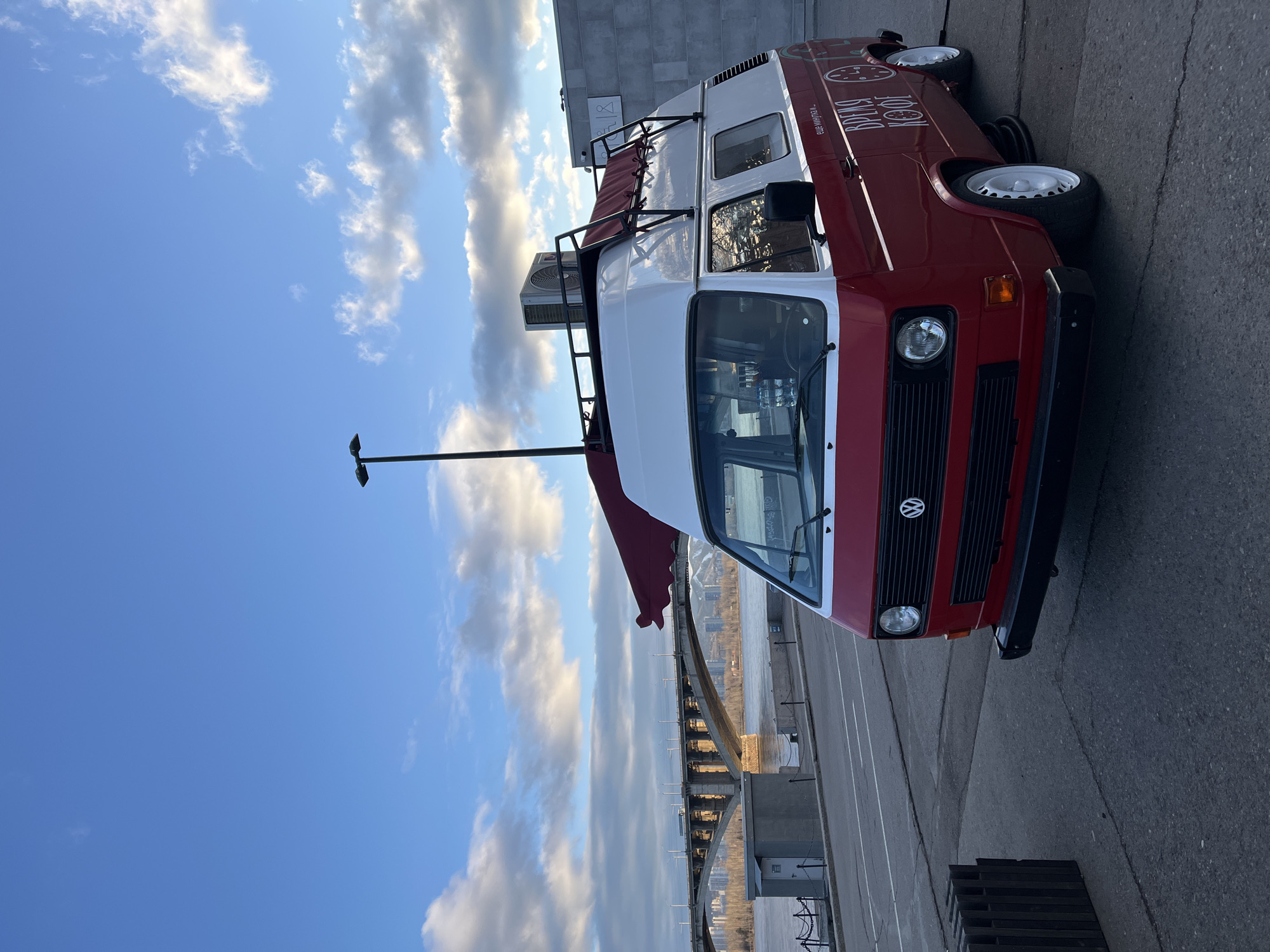 The image displays a two-tone red and white Volkswagen T3 van, likely serving as a mobile coffee shop, parked on a paved surface next to a river. The van features a red lower body and a white upper section, equipped with a roof rack holding an air conditioning unit. A red awning extends from the side, and the van rolls on distinctive white wheels. On the red side panel, white Cyrillic text is visible, reading "ВРЕМЯ КОФЕ" (Time Coffee) with additional smaller text below that appears to say "ещё минуточку..." (just a minute more...). A circular logo, possibly depicting a clock or coffee cup, is also present.
In the background, a wide river stretches, partially reflecting the sky, and is spanned by a prominent, multi-arch bridge. The sky above is a clear blue adorned with scattered white and grey cumulus clouds, illuminated by the warm light of late afternoon or early evening. The sun casts a gentle glow, suggesting pleasant weather. A tall, dark pole with a light fixture stands near the front of the van. Behind the van, a modern building with grey, textured walls is partially visible. On the ground near the van's front, several wooden pallets are stacked.
The scene appears calm and peaceful, devoid of any people or discernible activity beyond the stationary van. The presence of a large river and bridge aligns with the provided location context of Krasnoyarsk, Russia.