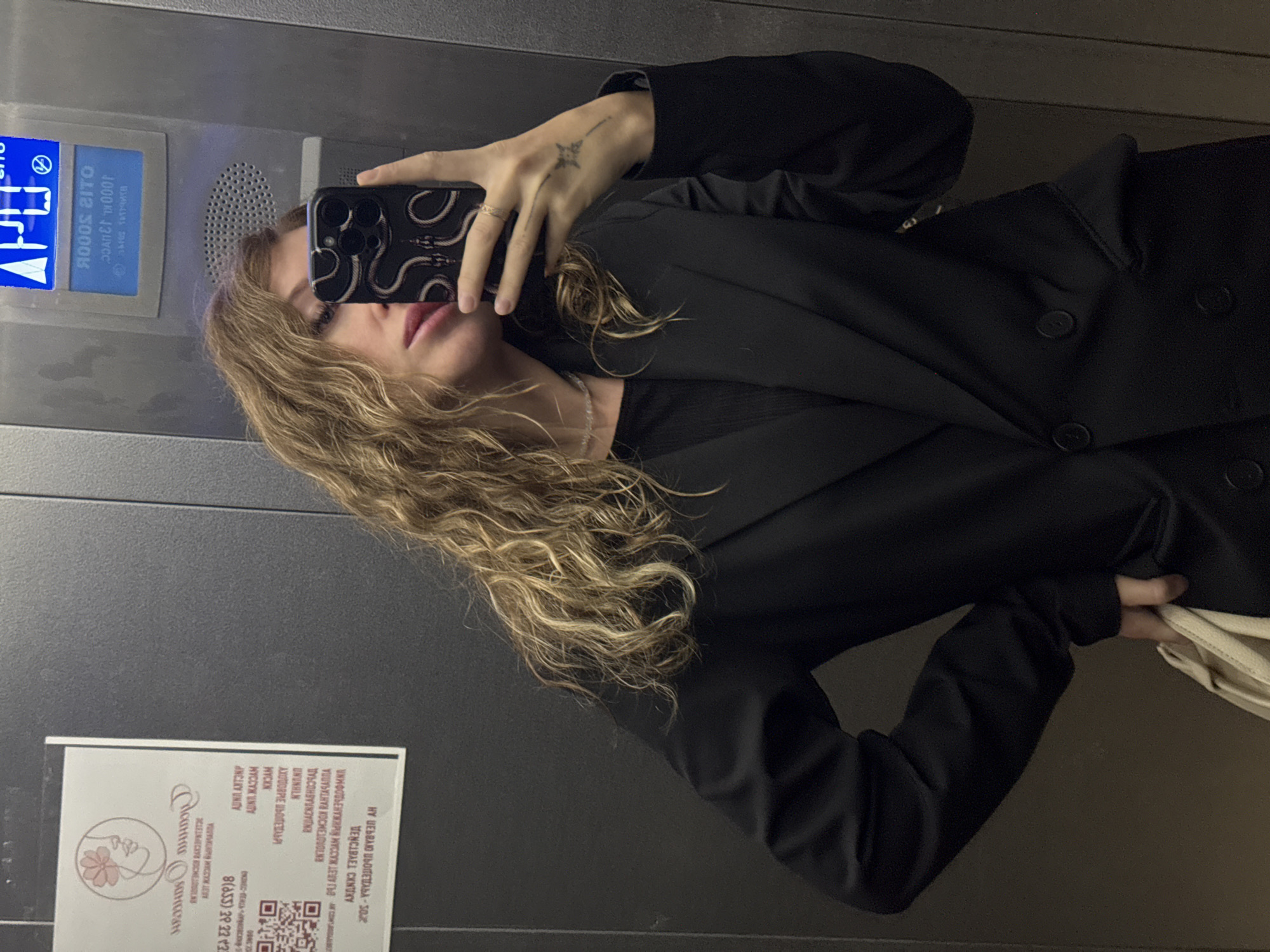 A woman is taking a selfie in an elevator. She is wearing a black blazer and a black top. She has long, wavy blonde hair. In her right hand, she is holding a smartphone with a snake-patterned case. On her left hand, she has a thin ring and a small tattoo of a star with lines extending from it. She is also holding a light-colored bag in her left hand. The elevator walls are made of dark grey metal. Above her, an elevator control panel is visible, with blue illuminated buttons and text in Cyrillic. A white sticker with a floral logo and text in Cyrillic is affixed to the left wall of the elevator. The image was taken in an elevator in Chaikovsky, Russia, as indicated by the Cyrillic text on the elevator panel and sticker.