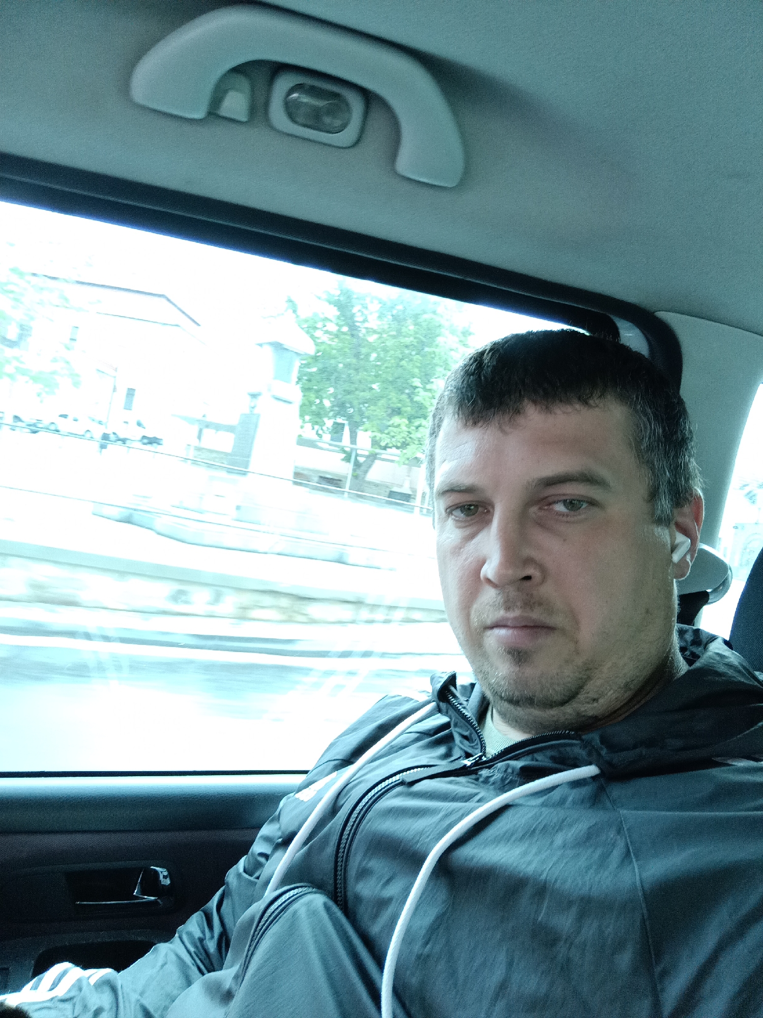A man with short, dark hair and light brown eyes is sitting in the back seat of a vehicle, looking directly at the camera. He is wearing a dark grey hooded jacket with white drawstrings. A white wireless earbud is visible in his right ear. The interior of the car is dimly lit, with a grey headliner and a grab handle visible above. The window on the left side of the vehicle shows a blurred, bright outdoor scene with buildings and trees. The scene is consistent with being inside a car in Dalovy, Czechia, likely during daytime.