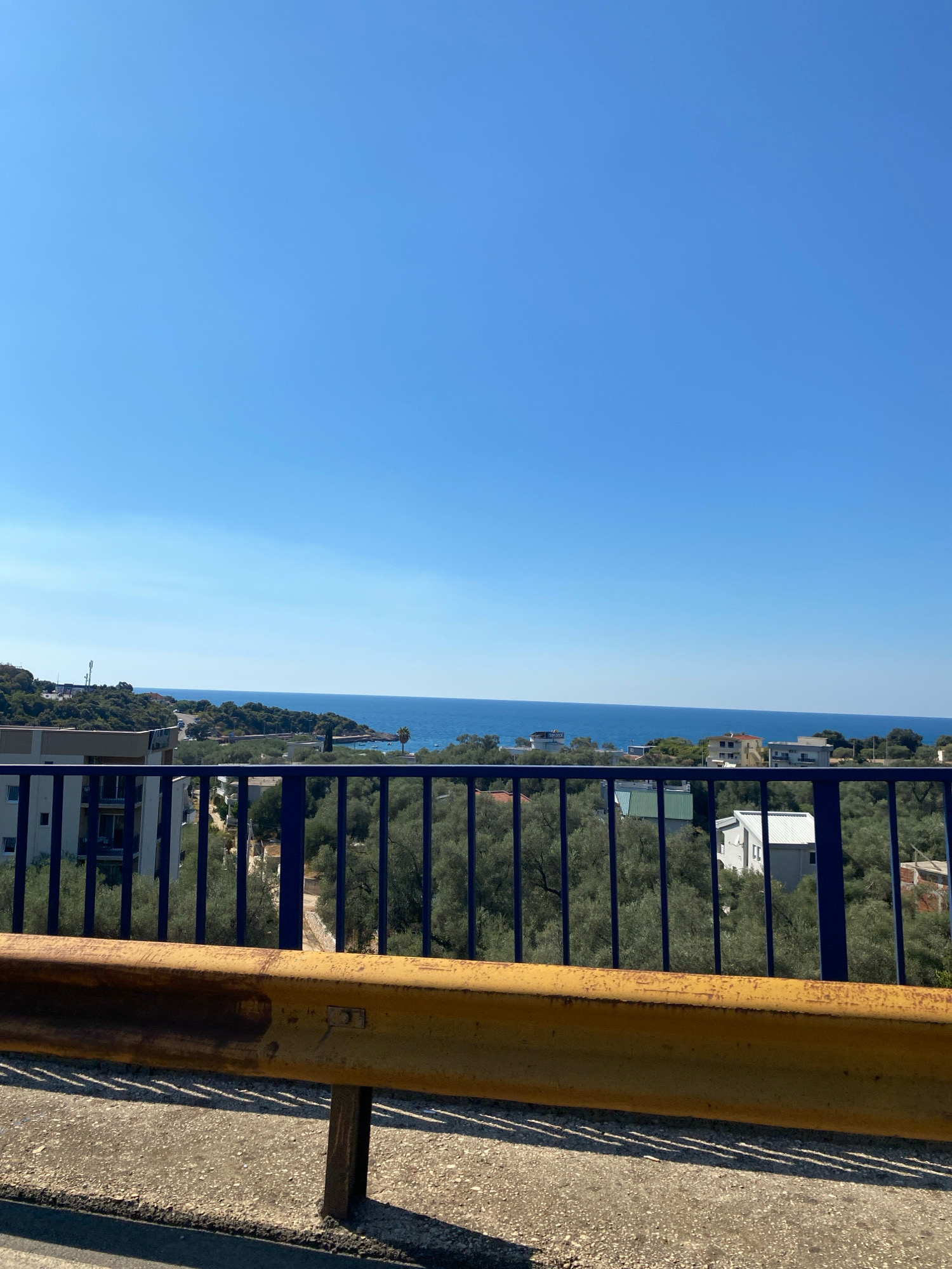 The image shows a view from a bridge overlooking the Adriatic Sea in Kunje, Montenegro. A yellow metal guardrail is in the foreground, casting shadows on the asphalt road. Beyond the guardrail, a blue metal fence separates the bridge from a landscape dotted with olive trees and several buildings, including a multi-story residential building on the left and smaller houses with red and white roofs further into the scene. The sea is visible in the middle ground, meeting a clear blue sky. The overall impression is a sunny day in a coastal town.