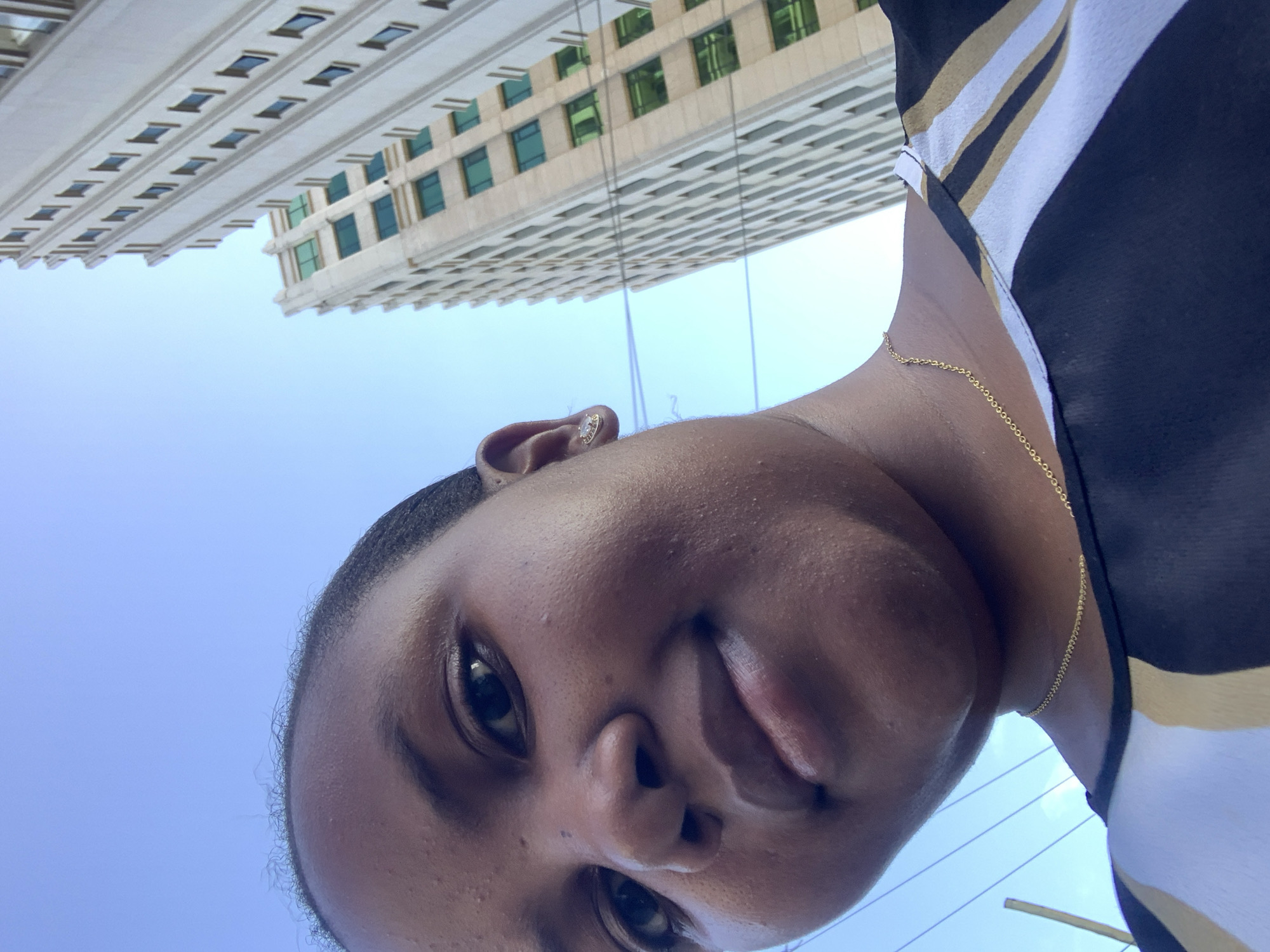 A close-up, upward-facing selfie photograph shows a person with dark skin, wearing a black, white, and gold striped top, and a delicate gold necklace. The person has short hair, and a small stud earring is visible in their left ear. Above the person, there's a view of a tall, light-colored building with many windows, set against a blue sky. Based on the architecture of the building, the location is likely in Dar es-Salaam, Tanzania, though this cannot be definitively confirmed from the image alone.