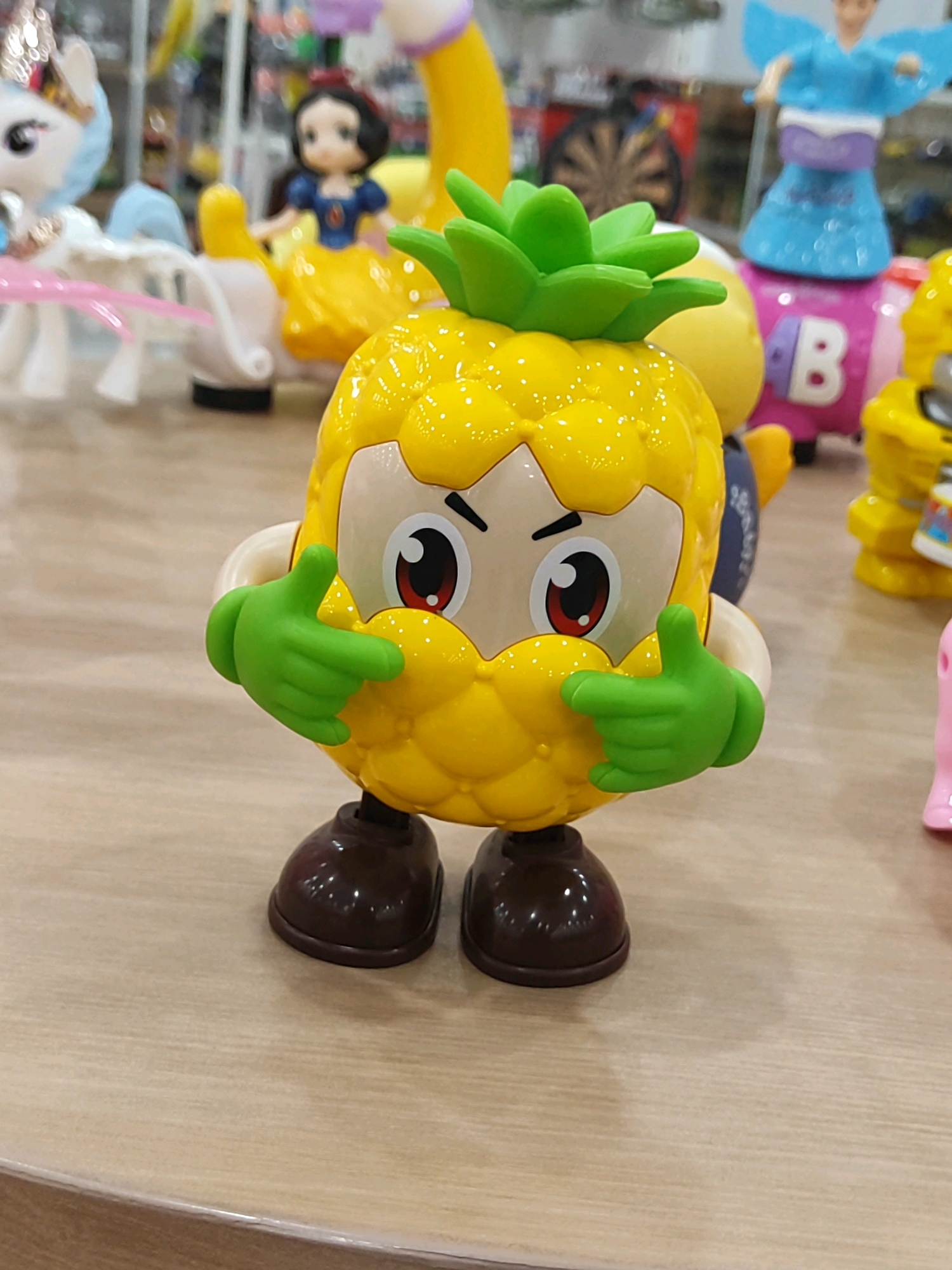 A toy pineapple character with green arms and a green leaf-like head stands on a wooden surface. The character has large red and black eyes and is positioned in the center of the frame. In the background, other toys are visible, including a white unicorn on the left and a figure in a blue dress on the right. The overall setting appears to be a store or display area. The image does not provide any information about the city of Sukowiryo, Indonesia.
