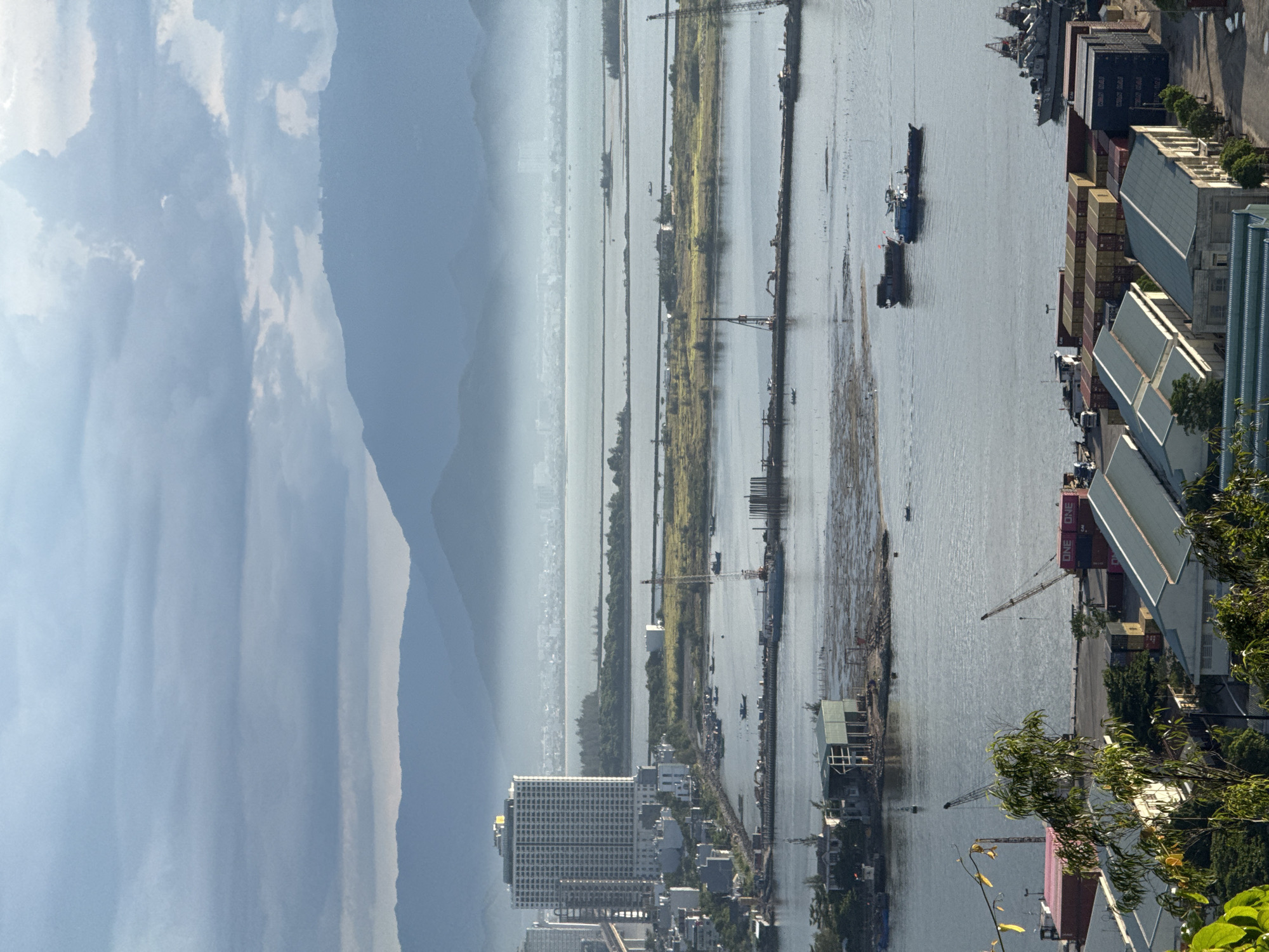 This is a high-angle, wide-shot photograph of a coastal urban and industrial area, likely in Da Nang, Vietnam, depicting a bay or river mouth with a prominent city skyline in the background. The foreground shows a densely packed industrial area with shipping containers stacked high, several modern industrial buildings with grey, sloped roofs, and trees. Two construction cranes are visible in the mid-ground, one red and white, and another yellow. A narrow walkway or pier extends into the water, with construction equipment and pilings. Several boats and barges are present in the water, including two larger vessels in the middle distance.
The mid-ground features a body of water, with a thin strip of land or artificial island running horizontally across it, dotted with vegetation and a few buildings. A large, multi-story building with a distinctive gold-colored roof, possibly a hotel, dominates the city skyline to the left of the frame. Beyond the city, a hazy, mountainous landscape forms the background, with layers of mountains receding into the distance. The sky is overcast with a mix of heavy grey clouds and patches of brighter, lighter sky, suggesting either early morning or late afternoon light. The overall impression is one of a busy, working port or industrial waterfront.