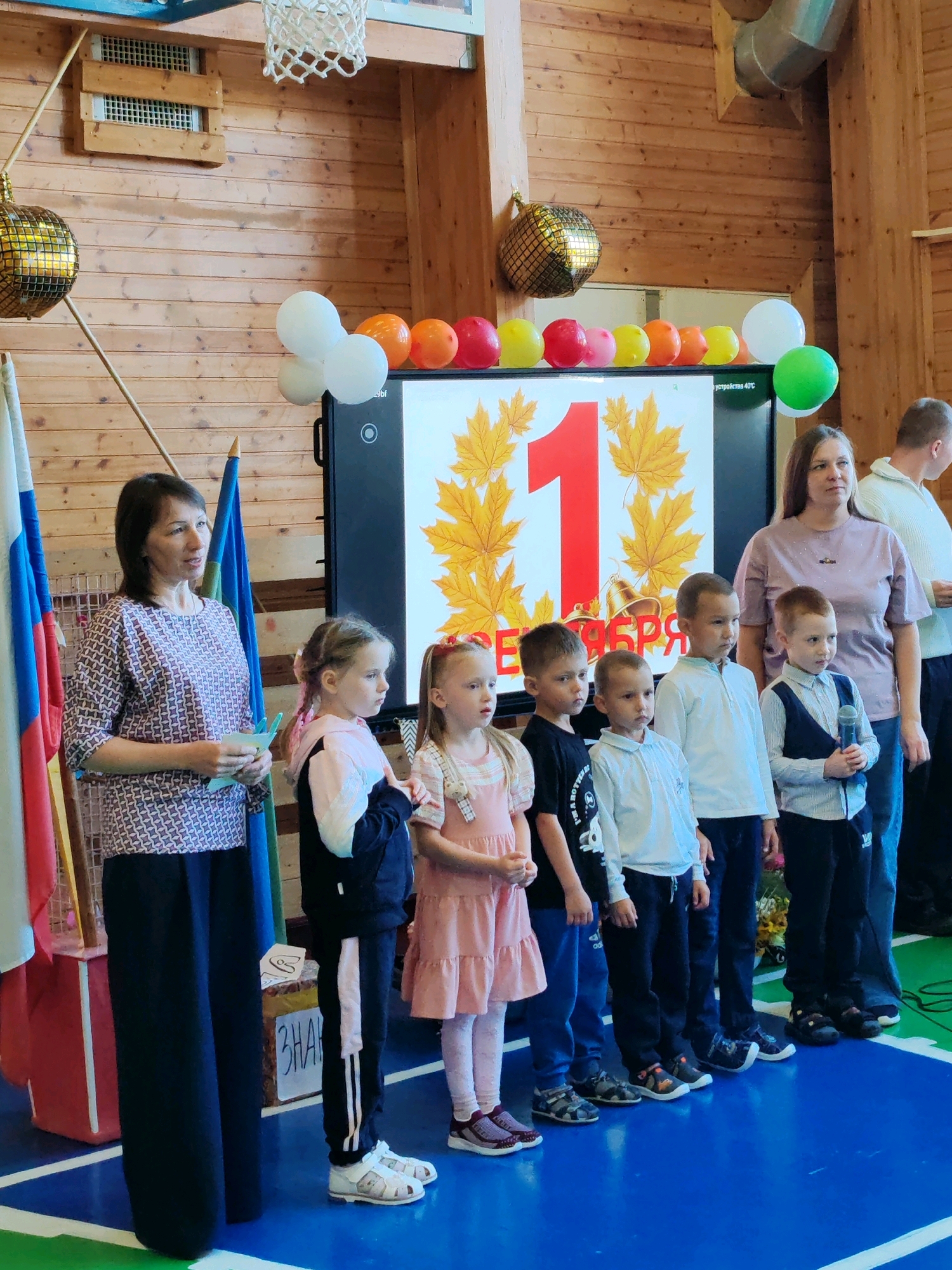 In an indoor gymnasium, a group of people, primarily children and two women, stand in front of a large screen displaying the number "1" surrounded by autumn leaves and bells. The screen is positioned on a wooden wall, and a row of colorful balloons, including white, orange, yellow, and green, are strung above it. A disco ball hangs from the ceiling on the left side of the image. The woman on the left, wearing a patterned blouse and dark pants, holds a piece of paper. A group of six children, varying in age, stand in a line from left to right, some looking at the screen, others to the side. The children are dressed in casual attire, with one boy holding a microphone. To the right of the children, another woman in a light purple t-shirt stands, also looking towards the screen. Behind the group, the wooden walls of the gymnasium are visible, along with a basketball hoop at the top left. The floor is a blue mat with green and white lines. Two Russian flags are positioned on the left side of the scene.