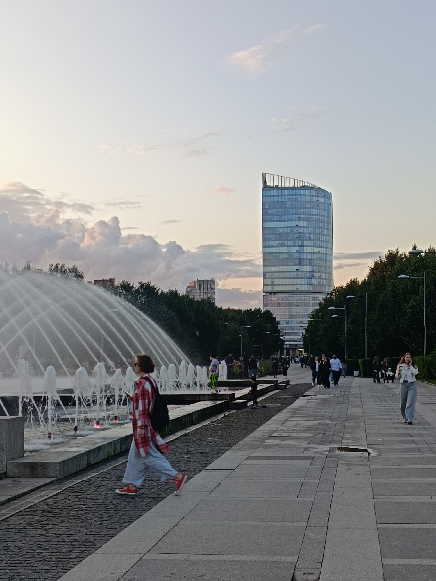 The image shows a wide paved walkway adjacent to a large fountain. On the left, the fountain sprays water in a wide arc. On the right side of the walkway, several people are walking, with a tall, modern glass skyscraper in the background. A woman in a red and white checkered shirt and light blue pants walks across the foreground, looking at her phone. The sky is overcast with soft clouds. The overall scene appears to be a public park or plaza in a city.