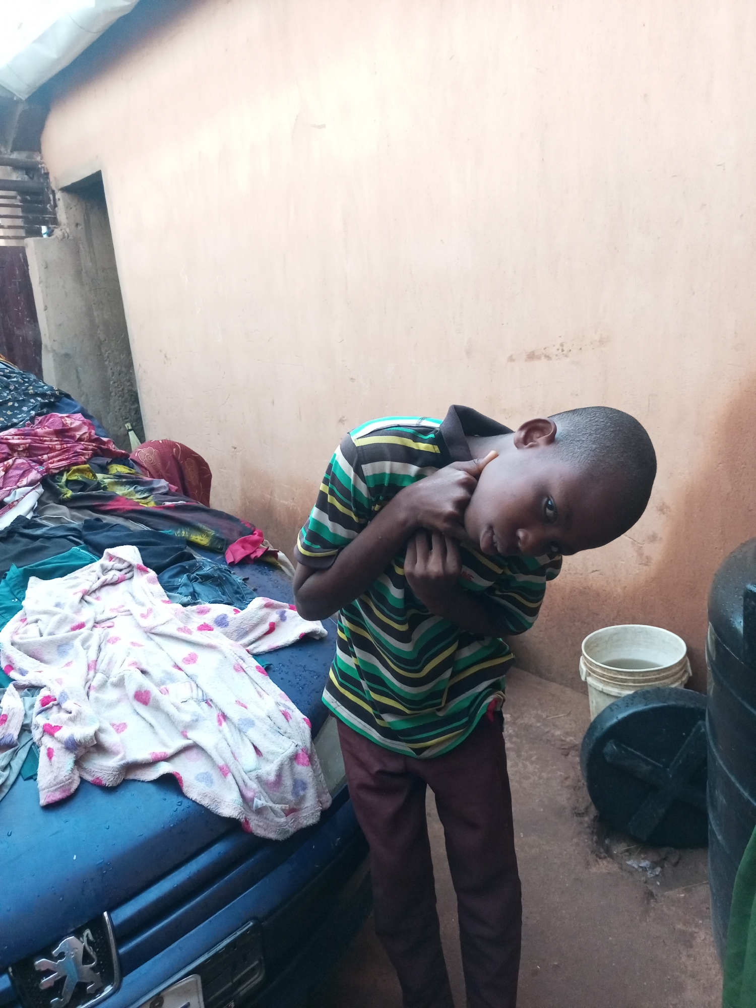 In Gombe, Nigeria, a young boy stands in an outdoor area next to a blue car and a wall. He is wearing a striped t-shirt and dark pants, and he is holding his hands to his neck. In front of him, various articles of clothing are spread out on the car, some of which appear to be a child's bathrobe with hearts. Behind the boy, a bucket of water and a large black tank are visible. The environment suggests a residential area, possibly a courtyard or yard, with a partially visible building structure on the left side of the image. The ground is dirt and appears wet in places.