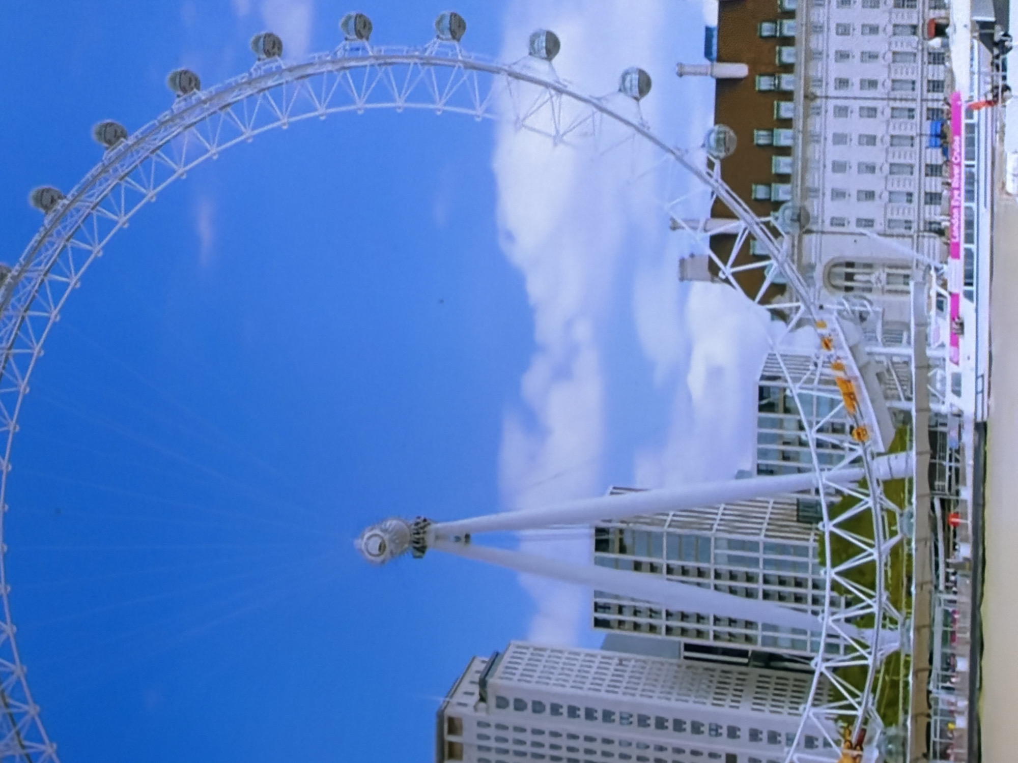 This image presents a prominent Ferris wheel, characterized by its extensive white lattice structure and numerous enclosed passenger capsules, curving upwards into a bright blue sky dotted with scattered white clouds. The wheel is supported by two large, white A-frame structures that converge at its hub. To the left of the supports, several tall, modern buildings with repeating window patterns are visible. To the right, a multi-story, classically designed building with a distinctive reddish-brown roof, light-colored facade, and evenly spaced windows stands. In the foreground, a body of water, likely a river, features several boats. One prominent white boat with a horizontal pink stripe clearly displays the text "London Eye River Cruise." The scene is set under natural daylight. No distinctly discernible human figures are present, though vague shapes consistent with people are visible on the boat. The described location for this scene is the city of Maidstone, United Kingdom.