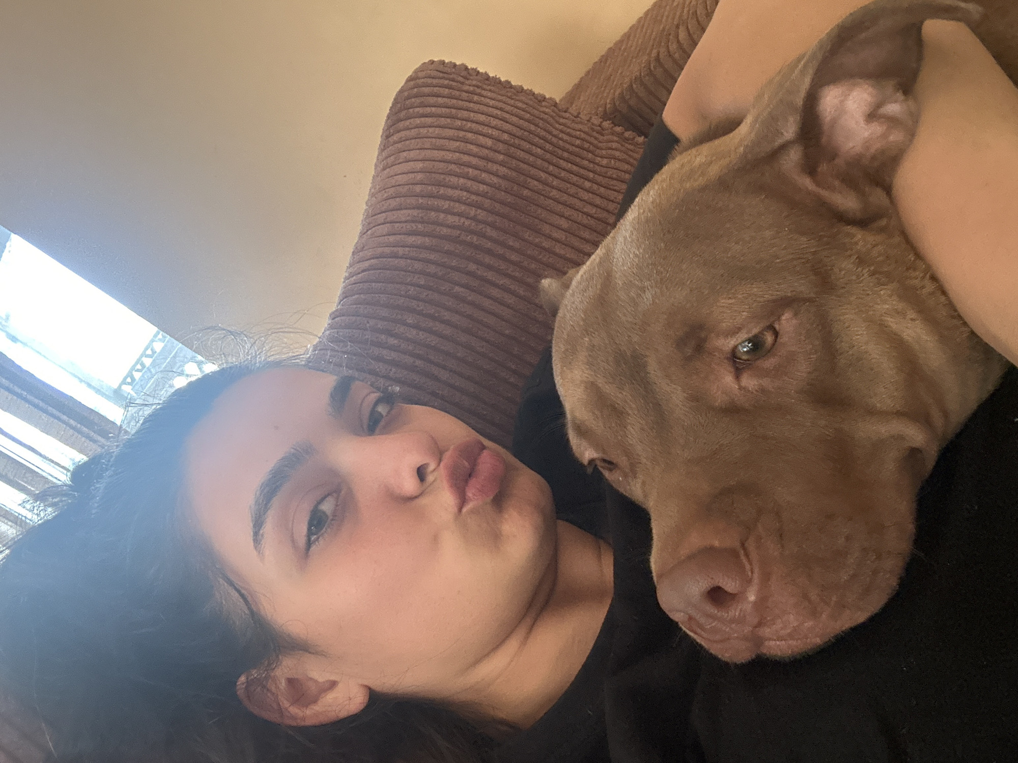 In Malevo, Bulgaria, a young woman with dark hair and striking eyebrows poses for a selfie, puckering her lips. She is wearing a black t-shirt and is nestled against a brown, corduroy armchair. Her head is tilted to the left, and her gaze is directed towards the camera. Beside her, a brown pit bull rests its head on her arm, its ear visible against her skin. The dog's eyes are open, and its nose is prominent in the frame. The background is softly blurred, with a suggestion of a window with blinds letting in natural light. The overall impression is one of a relaxed and intimate moment between the woman and her dog in a domestic setting.