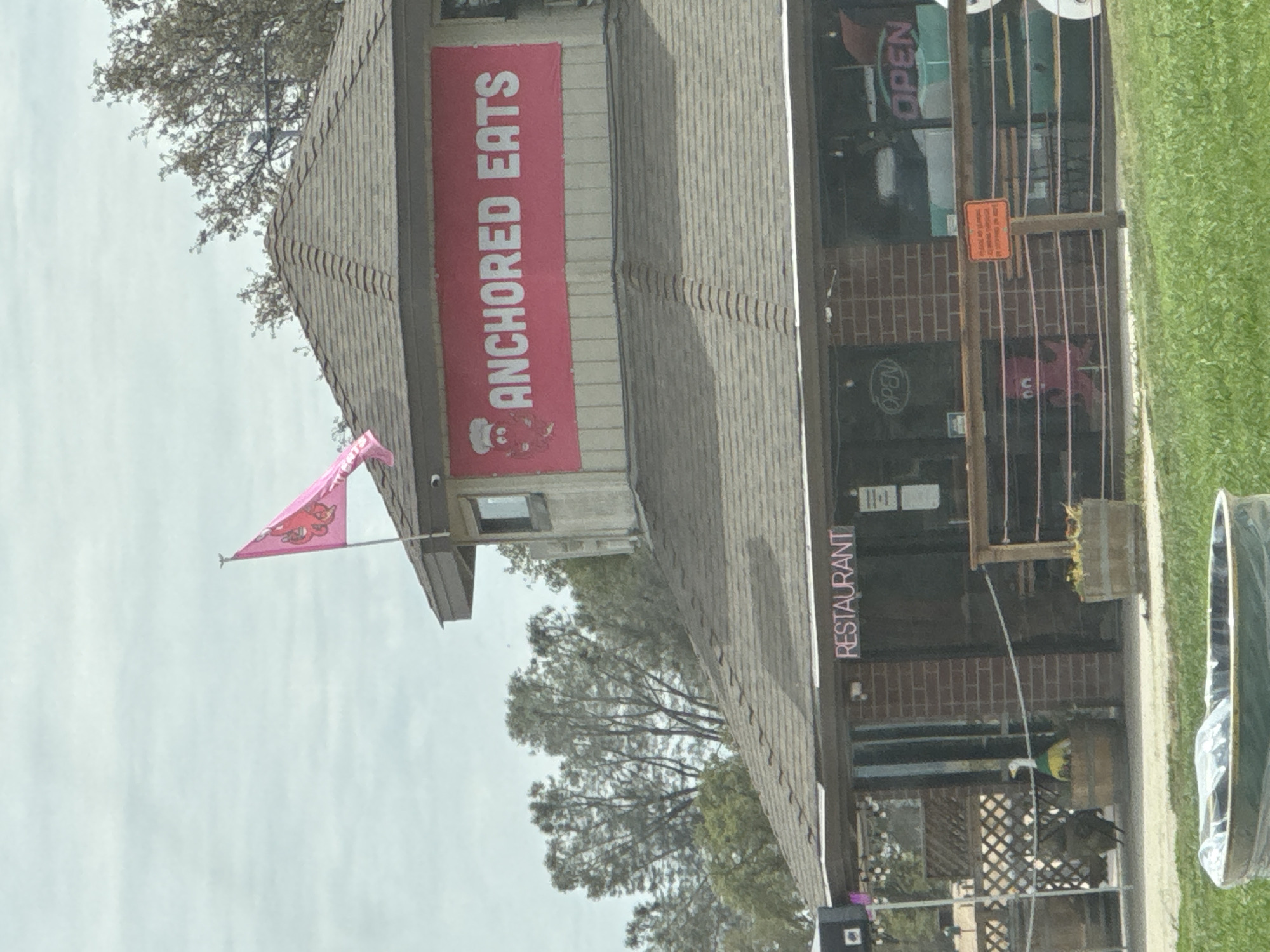 The image shows a restaurant called "Anchored Eats" in Folsom, United States. The restaurant has a red banner with white lettering that reads "ANCHORED EATS" and a pink flag with a cartoon octopus on top of the building. The restaurant also has a "RESTAURANT" sign above the entrance and "OPEN" signs visible in the windows. There are outdoor seating areas with wooden fences and barrel planters. In front of the restaurant, there are some decorative elements like a wooden barrel with a duck statue and another barrel with a pink octopus. A trash can is visible in the foreground. The sky is overcast.