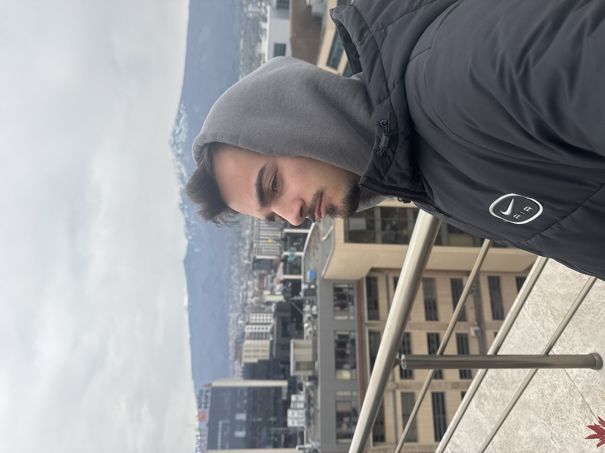 A man with a dark beard and hooded gray sweatshirt, wearing a black puffy jacket with a white Nike logo, stands on a balcony overlooking the city of Sofia, Bulgaria. The background includes numerous buildings and the Vitosha Mountain range, partially obscured by clouds. A metal railing is visible in the foreground. A red leaf lies in the lower right corner.