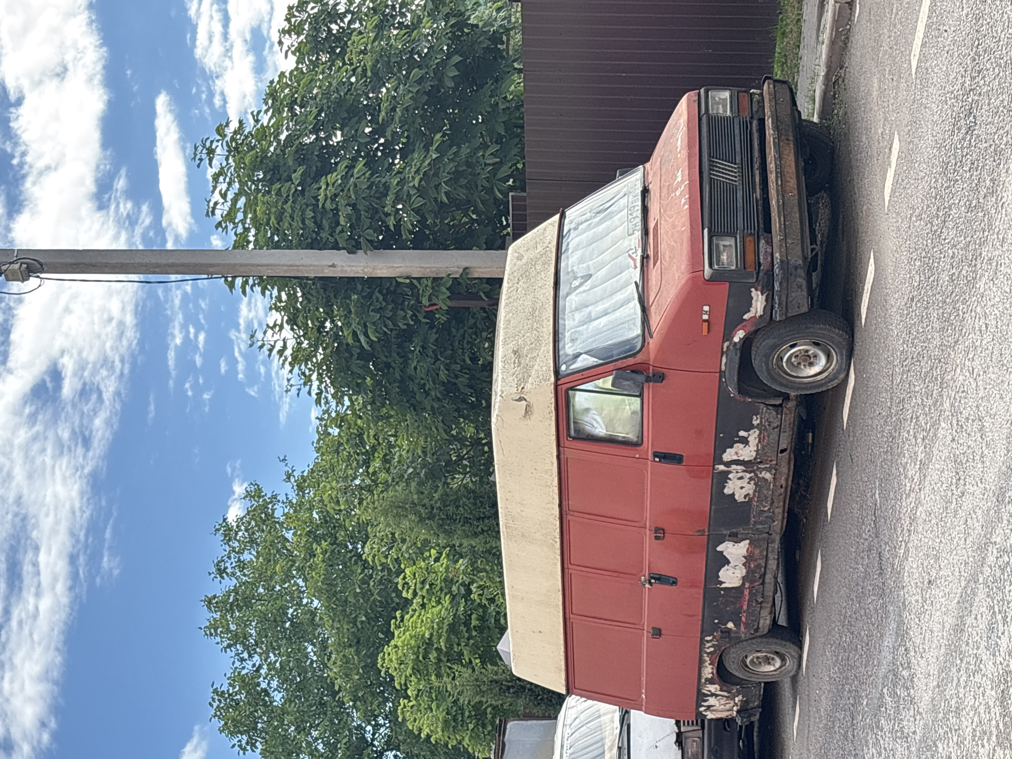 A red Fiat Ducato panel van with a beige raised roof is parked on the side of a paved road with white lane markings in Misaylovo, Russia. The van exhibits significant rust and paint damage along its lower body, with large patches of black primer showing through. It has a black grille and a damaged bumper. The van's windows are covered with a reflective sunshade. To the left of the van, a portion of another white vehicle is visible. Behind the van is a dark brown, corrugated metal fence, and beyond that, lush green trees and foliage. A concrete utility pole stands tall near the van. The sky above is blue with scattered white clouds. The scene appears to be an outdoor daytime setting.