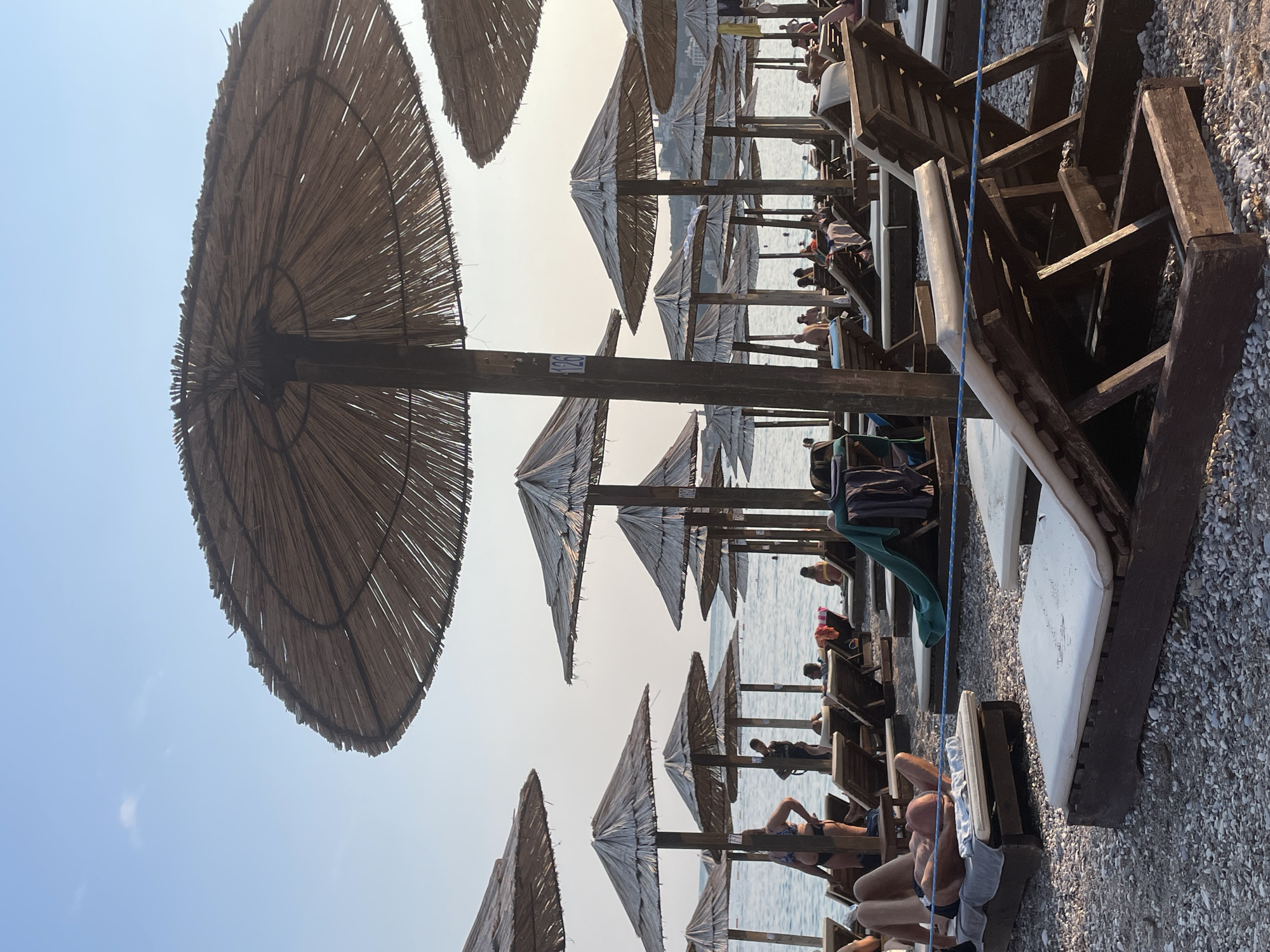 This media file depicts a beach scene in Budva, Montenegro, likely during the daytime. The image is oriented vertically, showing a row of straw umbrellas and wooden lounge chairs set up on a pebble beach. Several people are visible, some lounging on chairs, others perhaps walking or sitting near the water. The sea is visible in the background, with a hazy sky overhead, suggesting a warm and possibly sunny day, though the haze might indicate a slightly overcast sky or atmospheric conditions.
The subjects are primarily people and beach furniture. The straw umbrellas are a dominant visual element, providing shade. The lounge chairs appear to be made of dark wood and are arranged in neat rows. The beach itself is covered in small, light-colored pebbles.
The activity taking place is typical of a beach resort: people relaxing, sunbathing, and enjoying the seaside environment. The overall mood conveyed is one of leisure and vacation.
Notable details include the rustic appearance of the straw umbrellas and wooden furniture, which contributes to a natural, beachy aesthetic. The presence of multiple sets of umbrellas and chairs suggests a commercial establishment catering to beachgoers. There is a blue rope running through some of the wooden structures, possibly for structural support or as a decorative element. A small, white marker with the number "125" is visible on one of the wooden posts, likely indicating a specific spot or rental number.
The lighting suggests it is daytime, but the haze softens the sunlight. The location context of Budva, Montenegro, places this scene in a popular Adriatic coastal town known for its beaches.
