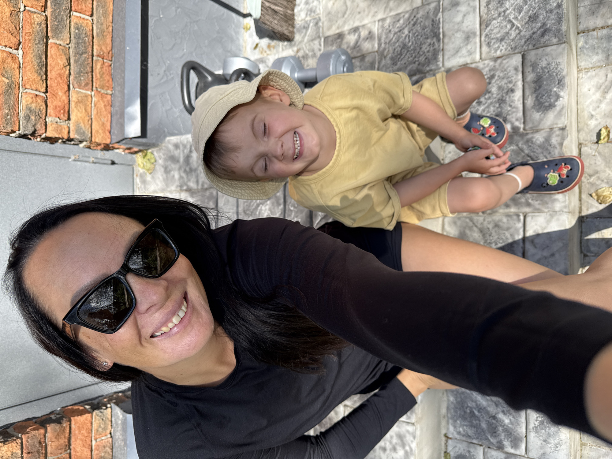 In this high-angle, eye-level selfie taken outdoors, a woman and a young child are the subjects. The woman, positioned on the left, is wearing a black long-sleeved shirt and sunglasses, smiling broadly. Her dark hair is visible, falling past her shoulders. To her right, the child is wearing a yellow t-shirt and a light-colored bucket hat, with their eyes closed and mouth open in a wide smile. The child is wearing dark blue clogs with red trim. They are both situated on a paved surface, likely a patio, with grey rectangular pavers. In the background, a brick wall is visible, and to the right of the child, a grey weight with a black handle and a broom with a light-colored handle are present. The scene suggests a sunny day, possibly in a residential area or park. The location is identified as Chaikovsky, Russia, though specific urban features are not prominent enough to pinpoint a precise spot within the city.