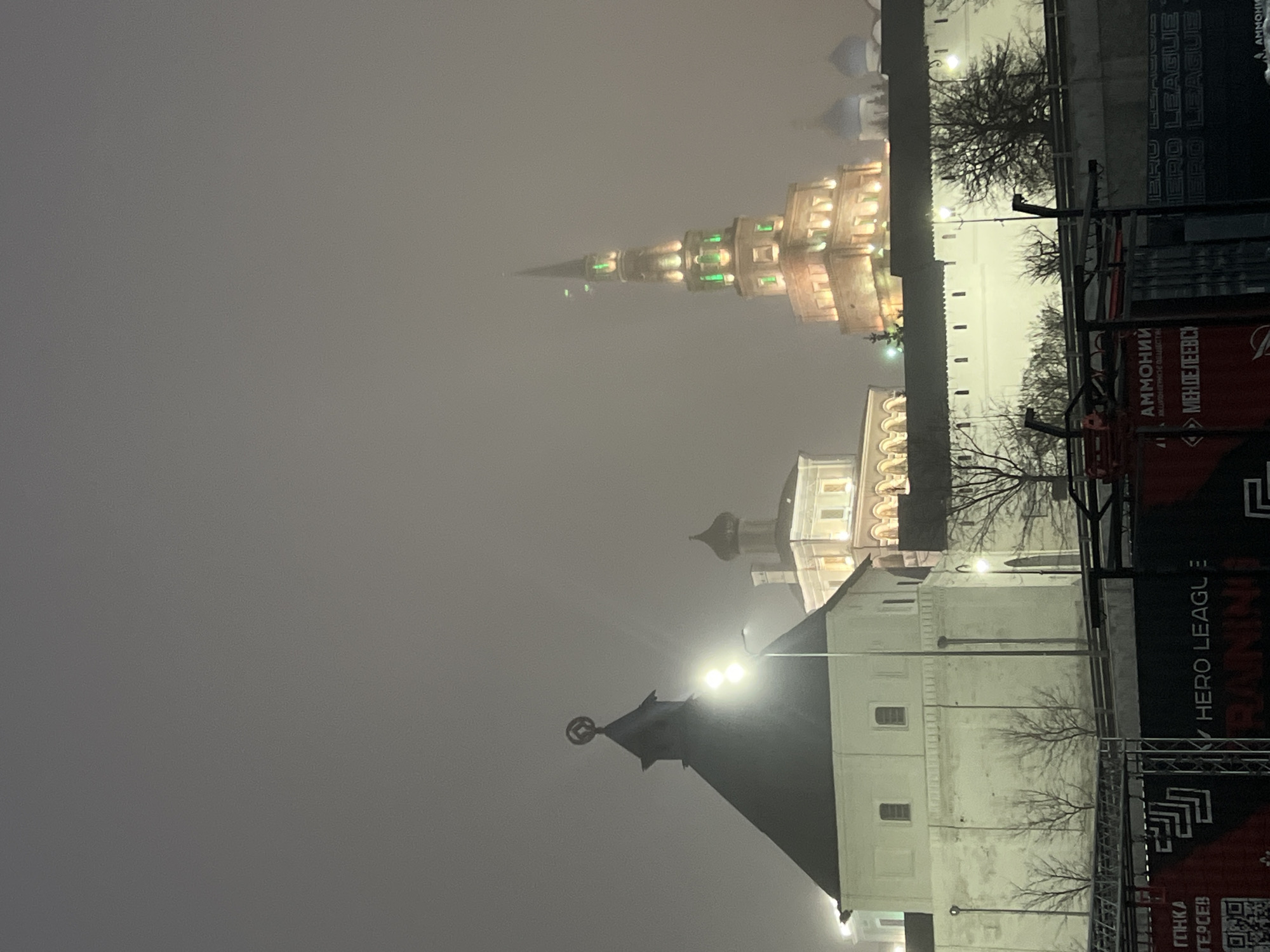 The image shows a low-angle view of Kazan Kremlin buildings under a foggy night sky. The tower of the Suyumbike Tower stands out with a star and its dark spire on top. In the background, the multi-tiered Spasskaya Tower is visible. Both towers are lit up. A large black banner is in the right foreground, obscuring parts of the image. The text on the banner includes "HERO LEAGUE" and "AMMO НИЙ МЕНДЕЛЕЕВС". Bare trees are visible between the buildings. The entire scene appears to be enveloped in a gray haze. The image is oriented vertically.