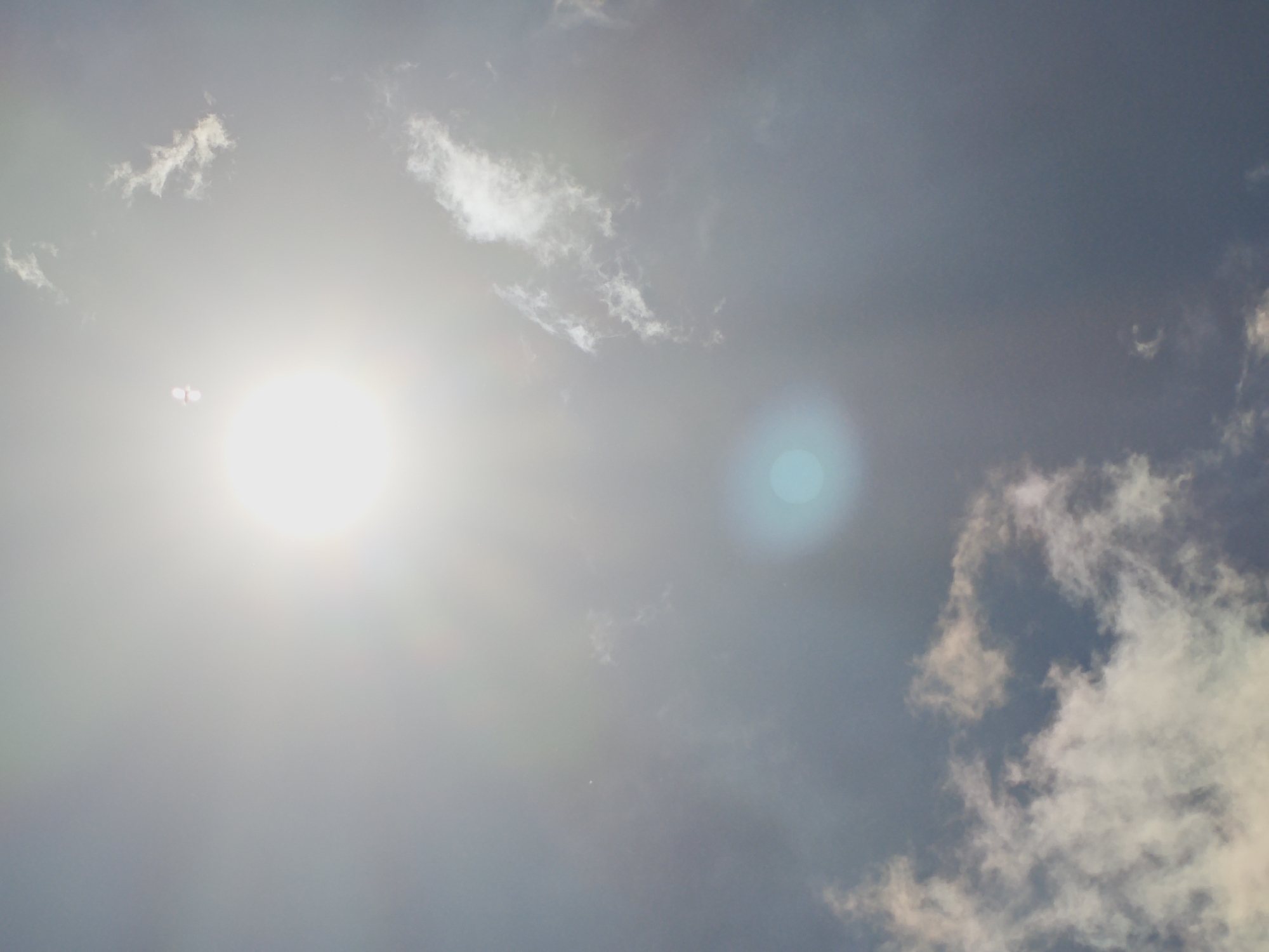 The image captures a bright sky with scattered clouds, indicating it is daytime. The sun is visible as a large, luminous orb on the left side of the frame, creating lens flare and washing out much of the surrounding area with its brilliance. A smaller, turquoise lens flare is also present towards the center-right of the image. Wispy white clouds are dispersed across the sky, some catching the sunlight and appearing brighter. The overall impression is that of a sunny day with some cloud cover. There is no indication of the city of Izgrev, Bulgaria, in the image.