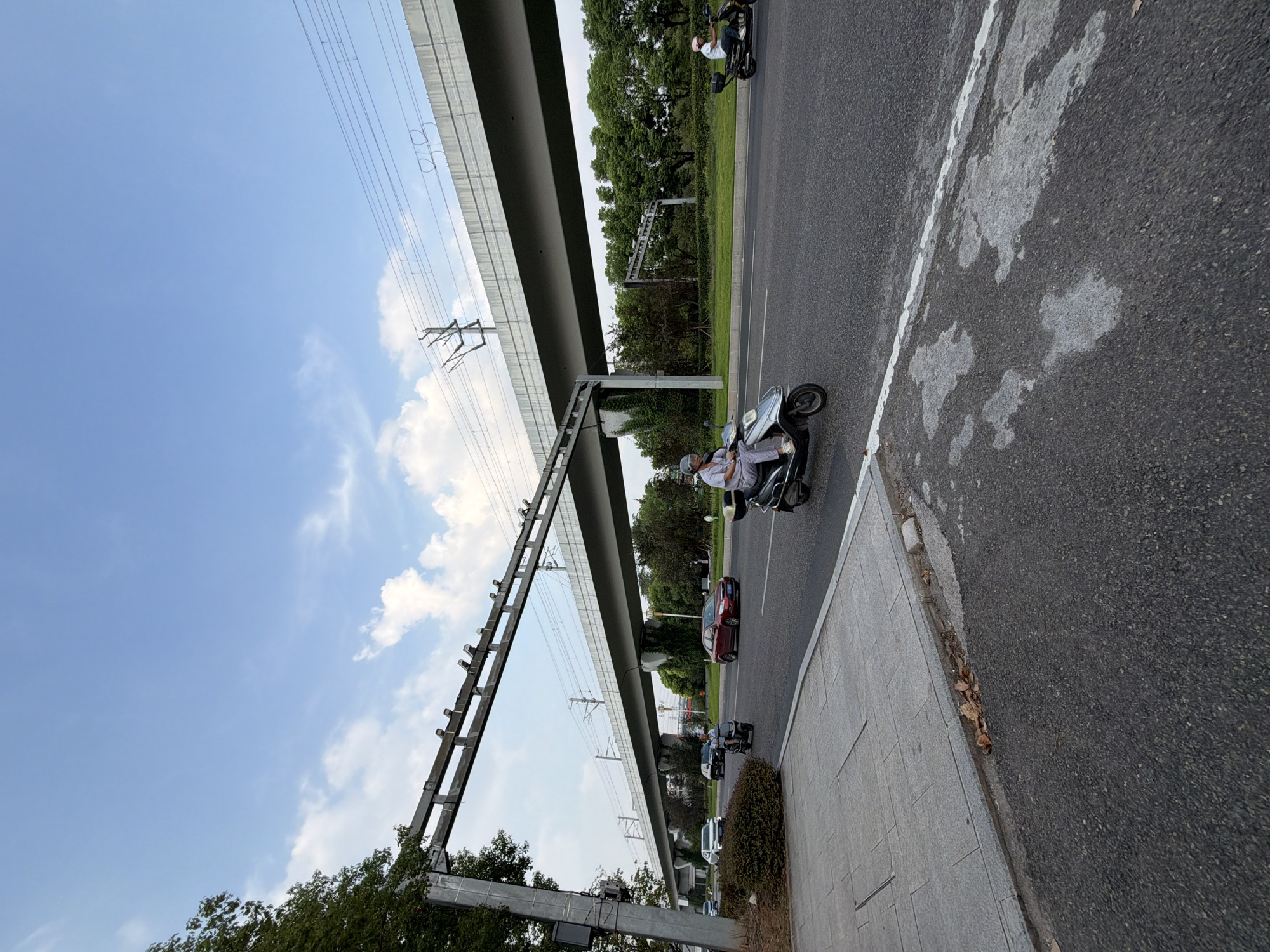 The image depicts a street scene in Jiangjin District, Chongqing, China, under a bright blue sky with scattered clouds. A multi-lane road runs horizontally across the frame, with vehicles present. A person on a silver scooter is in the foreground, moving towards the right. Behind them, a person on a black scooter is also traveling in the same direction. A red sedan is visible in the distance on the same road.
Overhead, a large elevated structure, likely part of an elevated railway or highway, spans the scene. This structure is supported by concrete pillars and has overhead wires running along its length, suggesting an electrified transport system. Trees and greenery line the sides of the road and the area beneath the elevated structure. To the left of the road, a paved sidewalk is visible, bordered by a gray concrete curb. The road surface is asphalt, with white lane markings. The overall impression is a modern urban environment with infrastructure designed for efficient transportation.