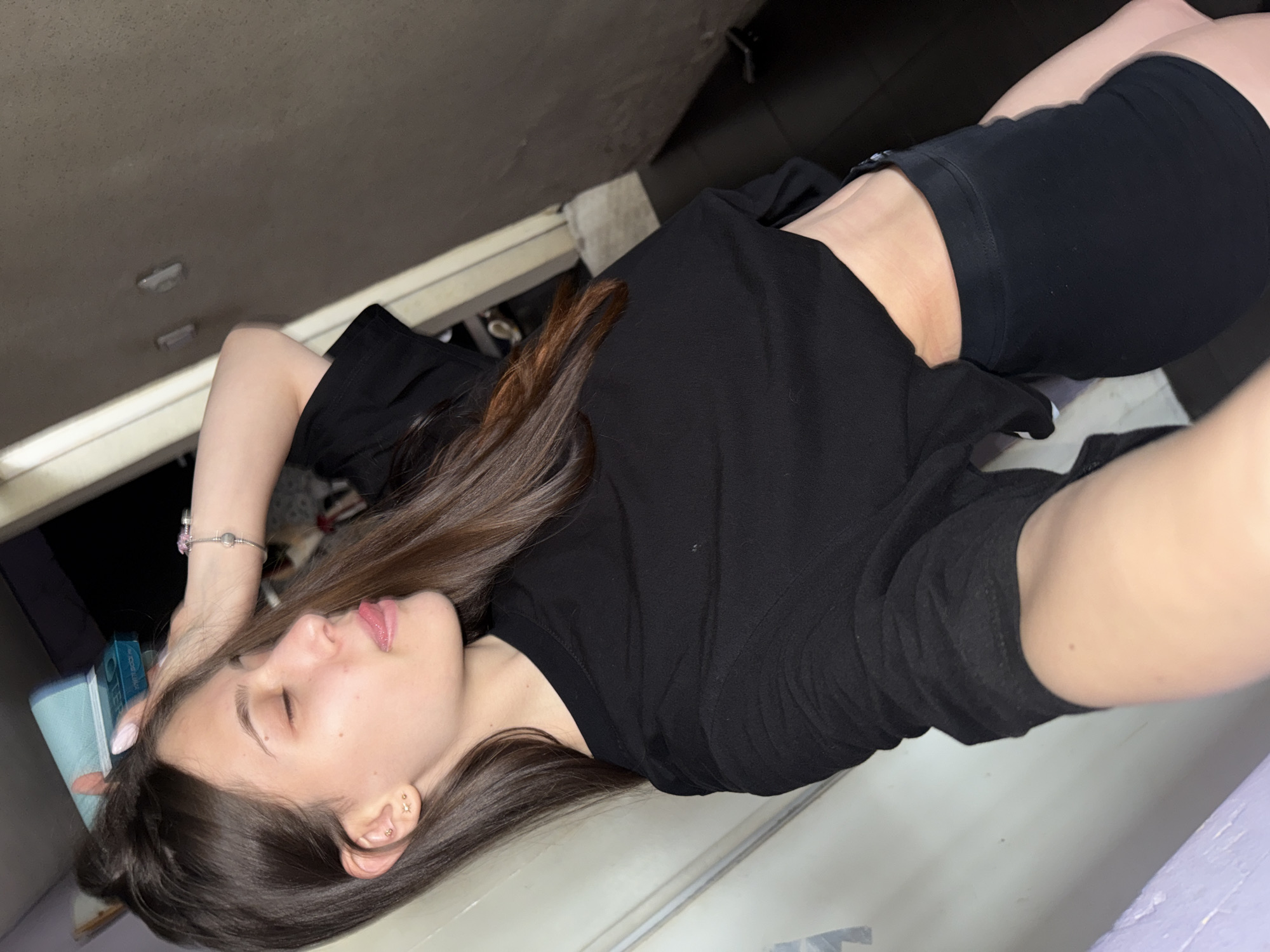 A young woman is lying down indoors. She is wearing a black t-shirt and black shorts. Her eyes are closed, and her brown hair is spread out. The location is not identifiable as Sofia, Bulgaria from the image.