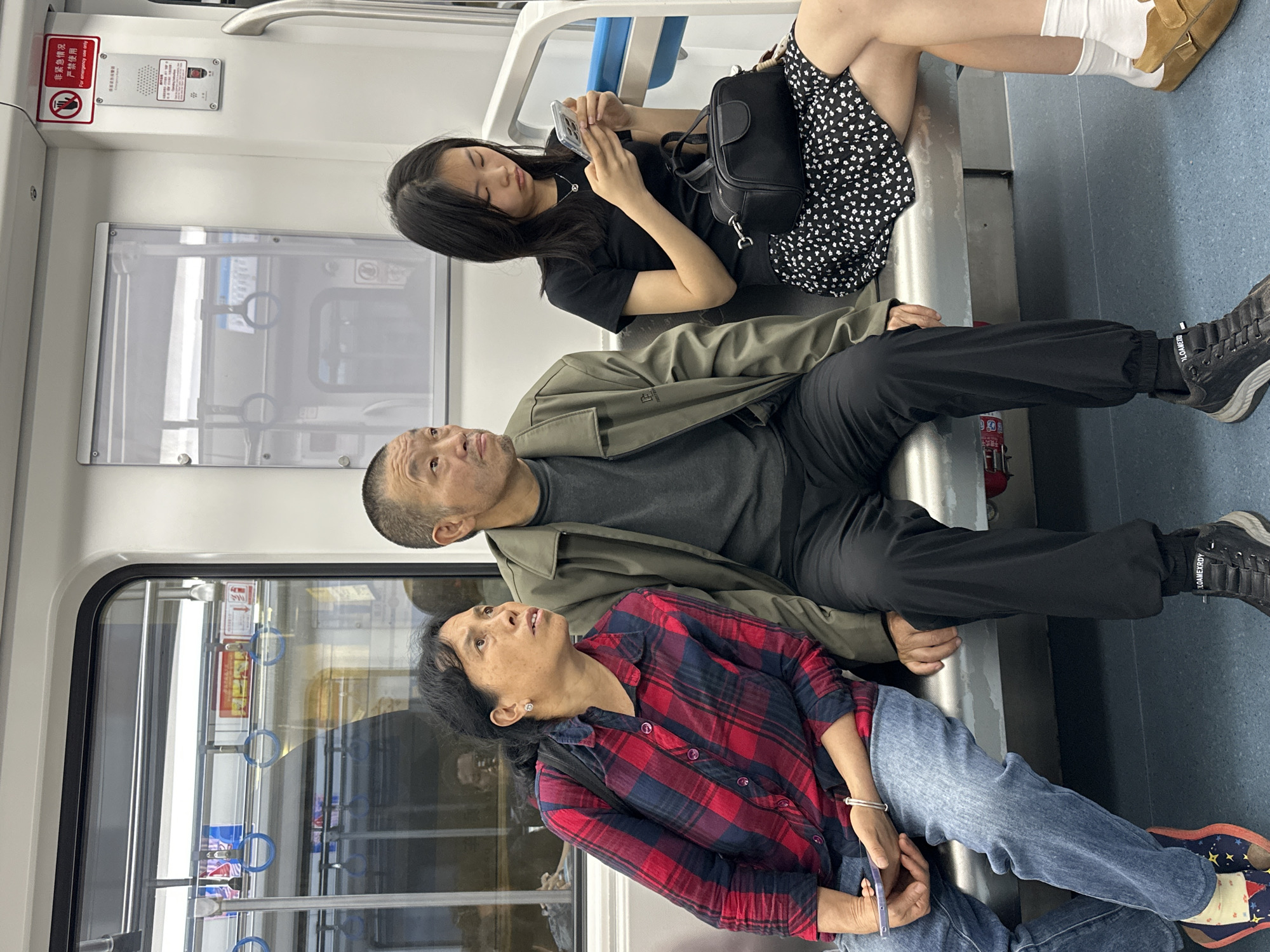 Three people are sitting on a bench in what appears to be a train or subway car. The person nearest the camera is an older woman with short black hair, wearing a red and blue plaid shirt and blue jeans. She is looking upwards. Next to her sits a middle-aged man in a gray shirt and a green jacket, also looking upwards. A young woman is seated farthest from the camera, wearing a black top and a black and white patterned skirt, looking at her phone. The interior of the train is visible through the windows, which are lined with metal handrails.