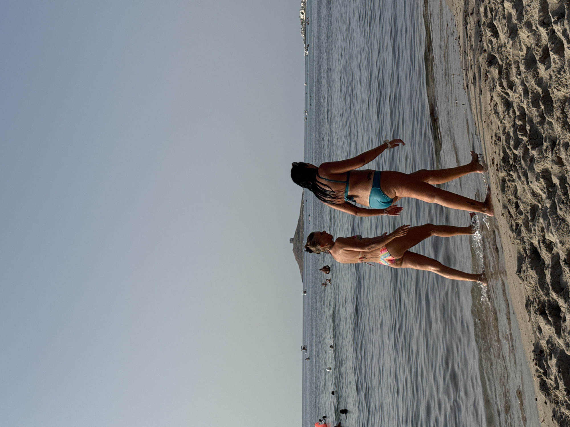 This is a medium shot of two women walking on a sandy beach in Isola delle Femmine, Italy. The woman on the left is wearing a colorful bikini and walking towards the sea. The woman on the right is wearing a blue bikini and walking parallel to the shoreline. The sea is calm and there are several people swimming and boating in the background. In the distance, there is a small island with a building on top. The sky is clear and blue. The sand is light brown and textured with small pebbles and shells.