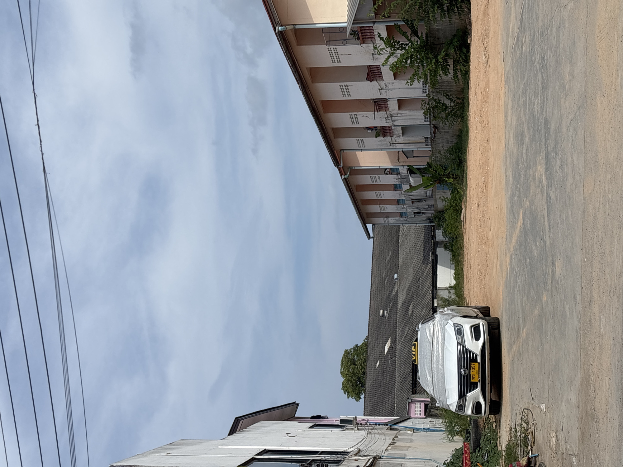 A white car, mostly covered in protective wrap, is parked on the side of a road in Choeng Thale, Thailand. The car has a yellow license plate with Thai script and the number 1357. Adjacent to the car, on the right, is a worn concrete road. A light-colored dirt shoulder runs alongside the concrete road. On the other side of the road, a multi-story building of peach and cream hues can be seen. Across the street from the car, there are low buildings and a tree in the background. Overhead, the sky is pale blue with light clouds. Electrical lines run parallel to the buildings.