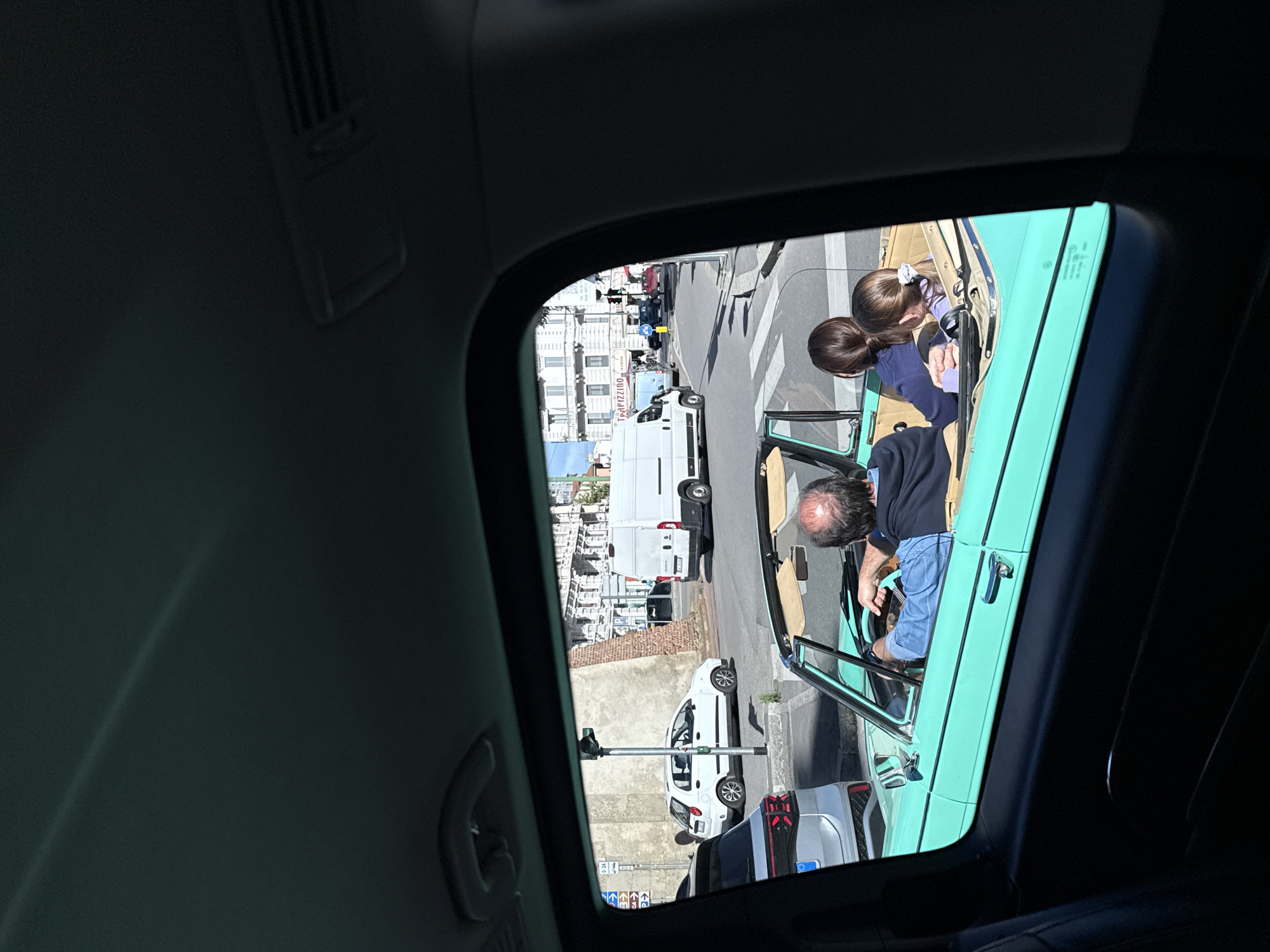 A view through the open sunroof of a car reveals a street scene in Milan, Italy. A vintage, open-top car with a turquoise exterior sits on the street. Inside, a man in a navy top and blue shorts is behind the wheel, while a young girl with brown hair, tied with a white ribbon, leans in towards the dashboard. The street beyond is lined with buildings, parked vehicles including a white van and a silver sedan, and a street lamp. Other pedestrians are partially visible in the distance. The perspective is slightly tilted.